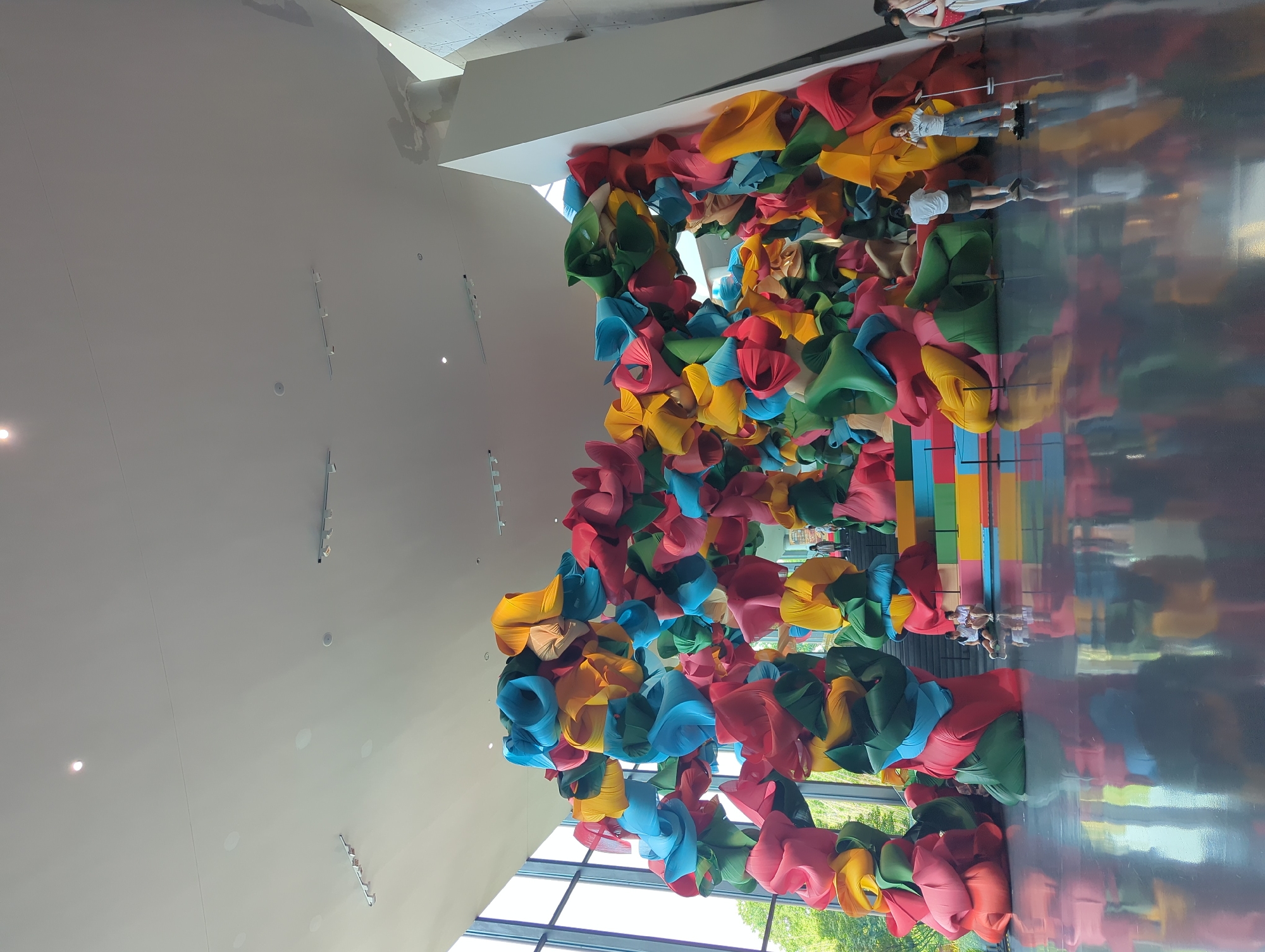 A large, colorful, abstract sculpture made of fabric forms dominates the center of a bright, modern indoor space, possibly a museum or gallery, located in Jinniu District, China. The sculpture is composed of many rounded, bulbous shapes in vibrant hues of red, yellow, blue, and green, twisted and layered to create an organic, cloud-like structure that arches over a set of multi-colored stairs. The floor is highly reflective, mirroring the sculpture and the ambient lighting. Several individuals are present: two people are seated on the stairs, appearing to be conversing; one person stands near the sculpture, looking at it; another person stands on a small platform near the right edge of the frame, holding what appears to be a phone; and in the far right, a person in a white outfit is partially visible. Large windows line the left side of the space, offering a glimpse of lush green trees and foliage outdoors. The ceiling is high and features recessed lighting fixtures. The overall impression is one of a dynamic and engaging artistic installation within a spacious, contemporary environment.