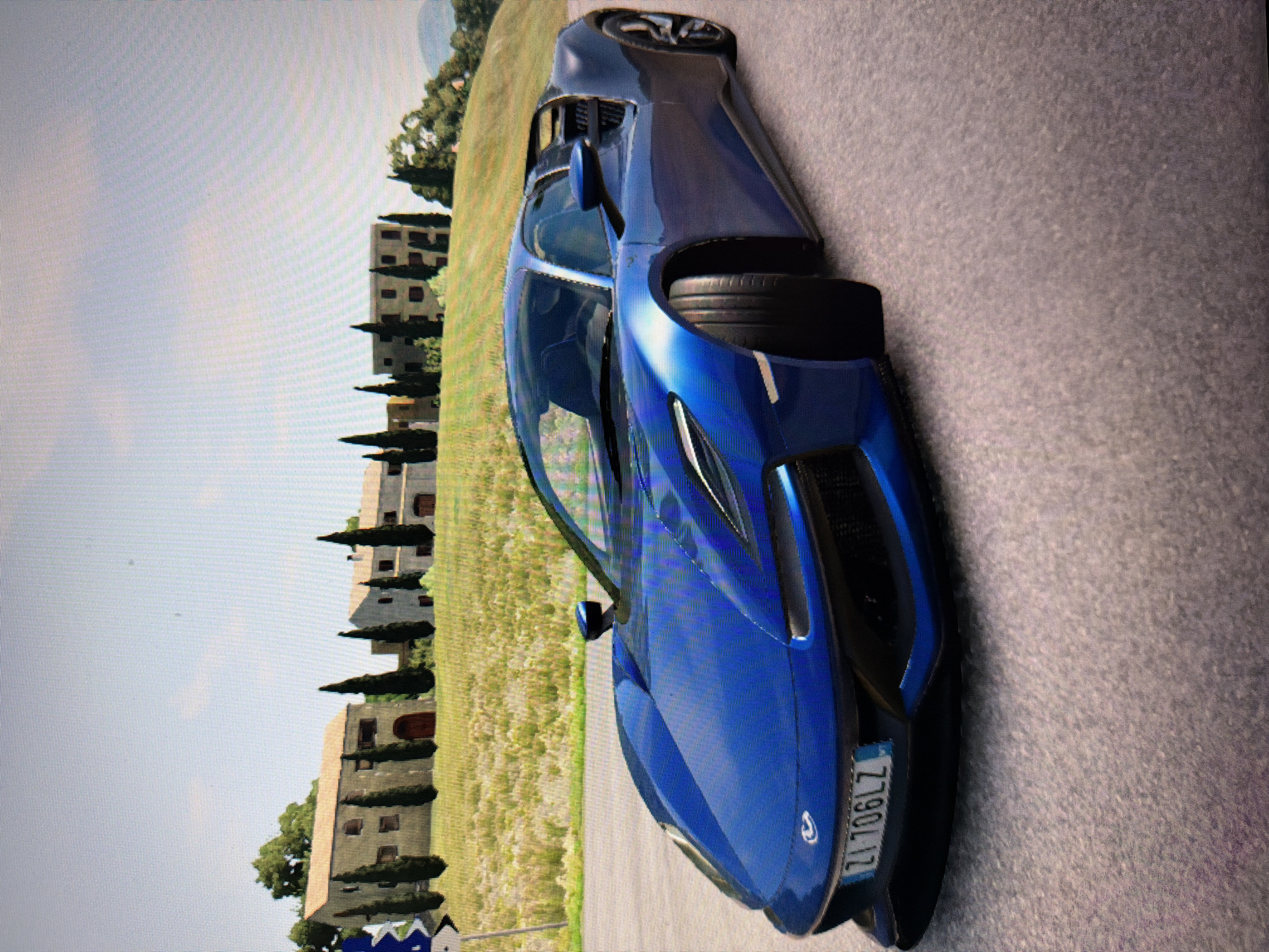 This image, initially oriented vertically, depicts a bright blue, sleek sports car parked on an asphalt road during daylight hours. The car, possibly an Acura NSX, is viewed from its front-left quarter, showcasing its aerodynamic design and glossy finish. On the front bumper, a license plate clearly displays "ZI 790 LZ".
The setting suggests a picturesque European countryside, likely Italy, due to the distinctive architecture and vegetation. In the background, a series of multi-story buildings with light-colored facades, arched doorways, and numerous small windows line a gentle slope. Tall, slender trees, resembling cypress trees, are interspersed among the buildings and dot the landscape. Beyond the buildings, a vibrant green field or vineyard extends towards the horizon. The sky above is a light blue with minimal cloud cover, indicating fair weather. There are no people visible, and the car appears stationary, suggesting a posed shot or a moment captured in a driving simulation. The pixelation and rendering style further indicate that this is likely a screenshot from a video game.