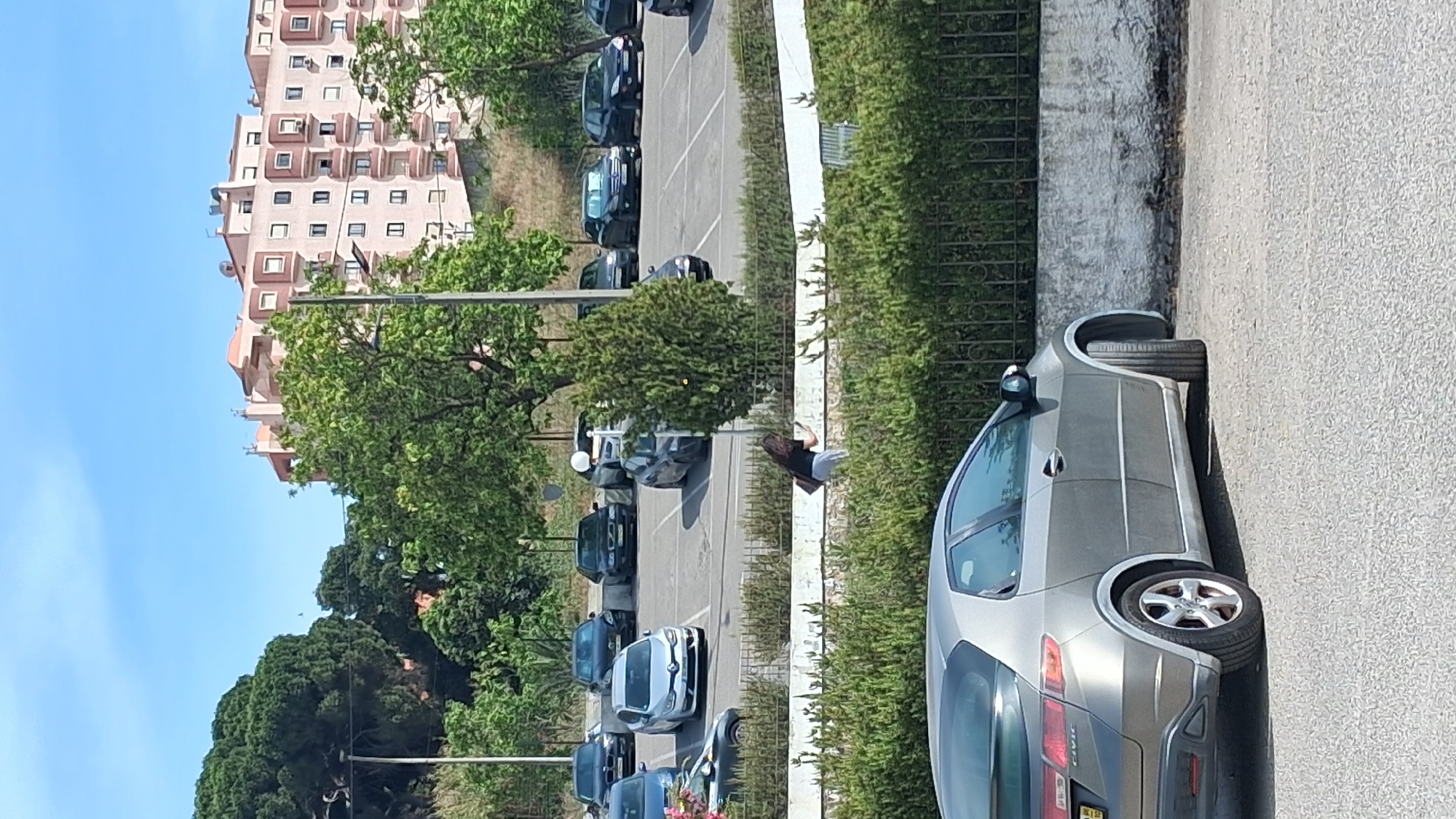 This is an outdoor, eye-level, medium shot photograph taken on a bright, sunny day. The scene depicts a parking lot with several vehicles and a person. In the foreground, a silver Honda Civic hatchback is parked on the right side of the frame, angled slightly towards the viewer. Behind it, a row of parked cars, including a white Renault Clio, a black BMW, and several other dark-colored sedans, are visible in a designated parking area. A woman with dark hair, wearing a black top and light-colored pants, is walking in the middle ground, holding a dark bag over her right shoulder and a phone in her right hand, appearing to be using it. She is walking between a hedge and the parking lot. In the background, a large, multi-story residential building with a pale pink facade and numerous balconies dominates the upper right portion of the image. Lush green trees, including a large pine tree on the left, and a smaller, dense evergreen tree in the middle ground, provide natural elements. The sky is a clear blue with a few wispy clouds. The overall environment suggests a residential area with a parking facility, possibly in the city of Setúbal, Portugal, given the architectural style of the building and the Mediterranean vegetation.