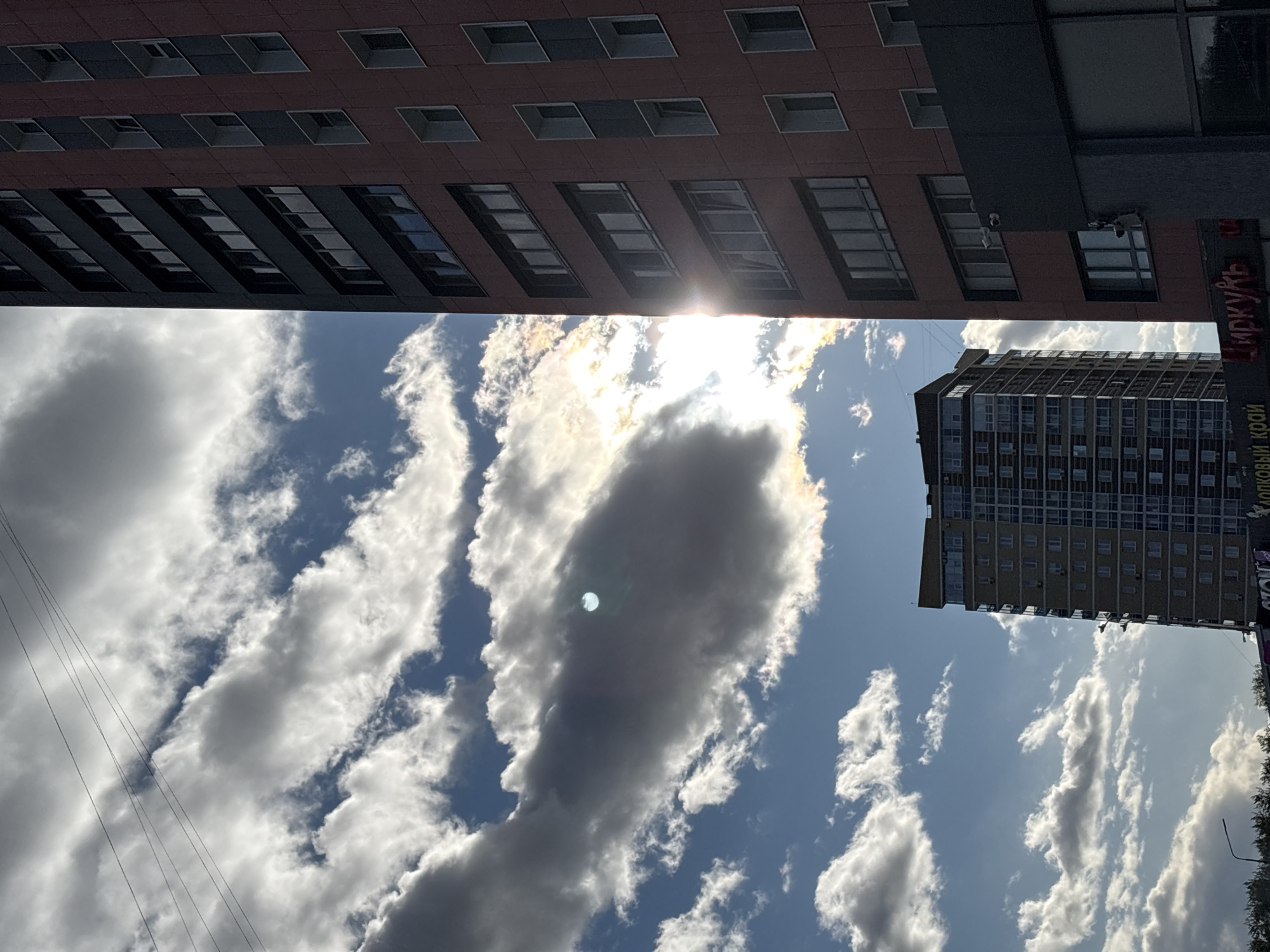 The image shows a view upwards in Perm, Russia, with two buildings framing a sky filled with clouds. The building on the top is a multi-story structure with rows of windows. The building on the right is also tall with a similar architectural style, featuring evenly spaced windows. The sky is blue with a scattering of puffy, white and gray clouds. The sun is shining brightly, partially obscured by the clouds, creating a glare in the center of the frame.