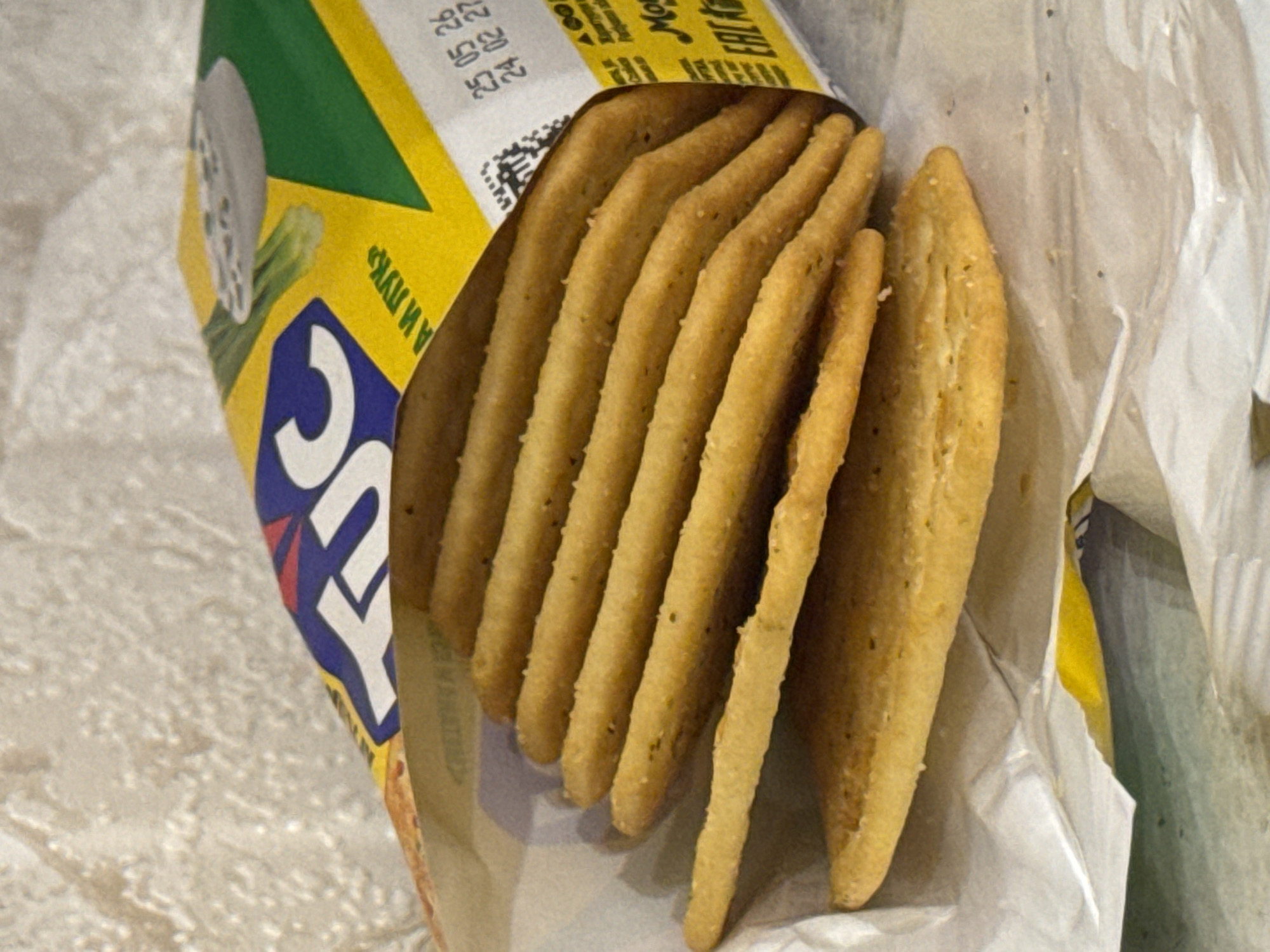 A close-up, eye-level, indoor shot captures a stack of golden-brown crackers spilling out of their packaging. The crackers, rectangular and thin, are layered unevenly, with the bottom ones forming a base and subsequent crackers angled upwards. Specks of seasoning are visible on their surfaces. The packaging, partially visible, features a yellow background with green and blue graphic elements, including the white lettering "TUC". A date stamp is visible on the packaging, reading "25 05 26" and "24 02 27". The scene appears to be a domestic setting, possibly a kitchen counter, with a textured, light-colored surface in the background, likely a tablecloth or placemat. The lighting is soft and natural, highlighting the texture and color of the crackers. The image does not contain any information to suggest a specific location in Saratov, Russia.