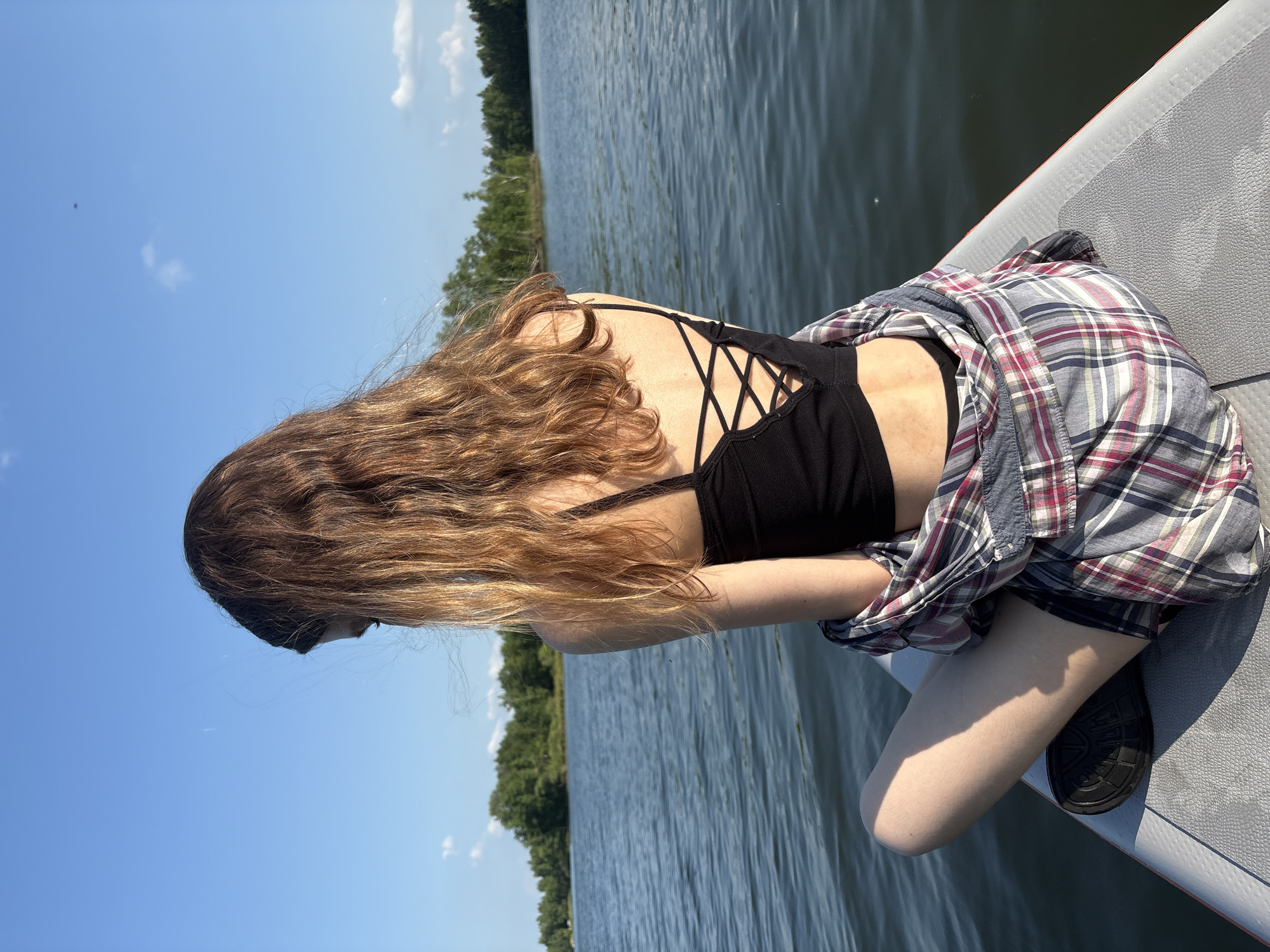A young woman with long, wavy brown hair is sitting on a paddleboard on a calm body of water, facing away from the camera. She is wearing a black criss-cross back tank top, a plaid shirt loosely draped around her shoulders, and dark shorts. Her legs are crossed, and she is looking down and to her left. The background shows a shoreline lined with green trees and a clear blue sky with a few wispy clouds. The water is rippling gently, reflecting the sky and surrounding foliage. The location is not specified in the image but is assumed to be in Razdolnoye, Russia.