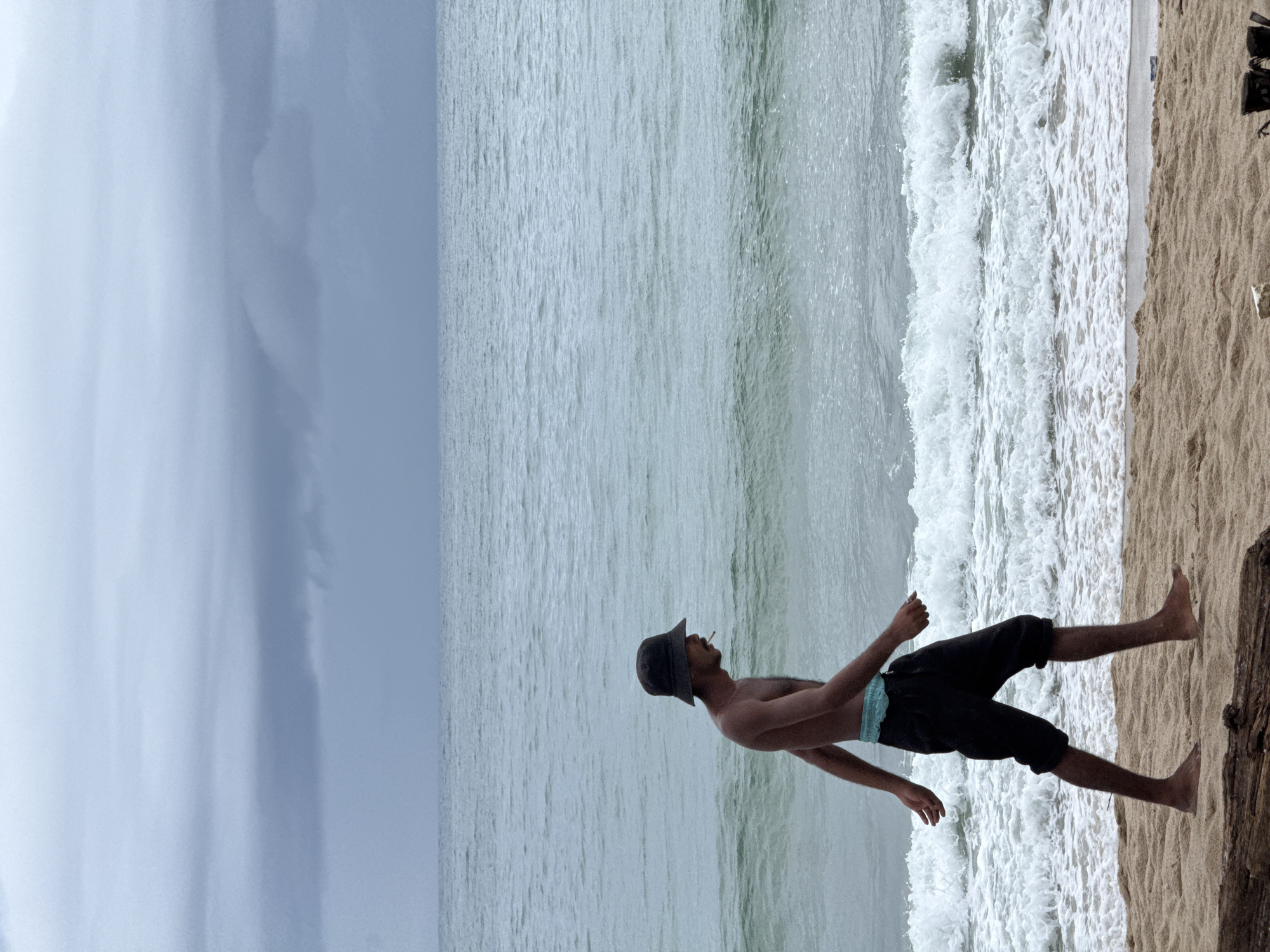 A man wearing a dark bucket hat and black shorts is walking on a sandy beach. He is bare-chested and has a cigarette in his mouth. The ocean is to his right, with waves crashing on the shore. The sky is cloudy and overcast. The scene appears to be in a coastal area, likely a beach in Thailand. The man is walking from left to right in the frame. The sand is a light brown color, and there is a piece of driftwood in the foreground. The water is a light blue-green color. The overall atmosphere is somber due to the overcast sky.