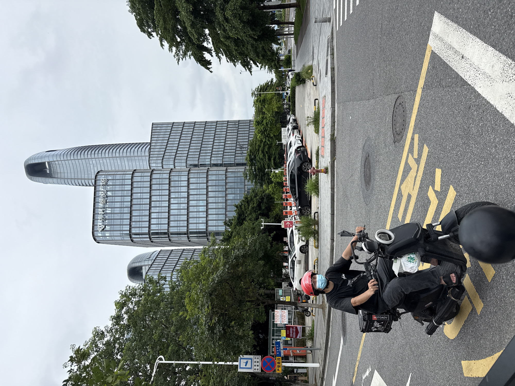 In Haizhu District, China, a person wearing a red helmet and a blue face mask rides an electric scooter on an asphalt road marked with yellow lines. The scooter has a black cargo box labeled "MOTOMAX" and a white plastic bag hanging from the front. The person is dressed in a black t-shirt and dark pants. In the background, several modern glass-covered buildings stand under a cloudy sky, with trees lining the street. A parking lot with various cars is visible between the road and the buildings.