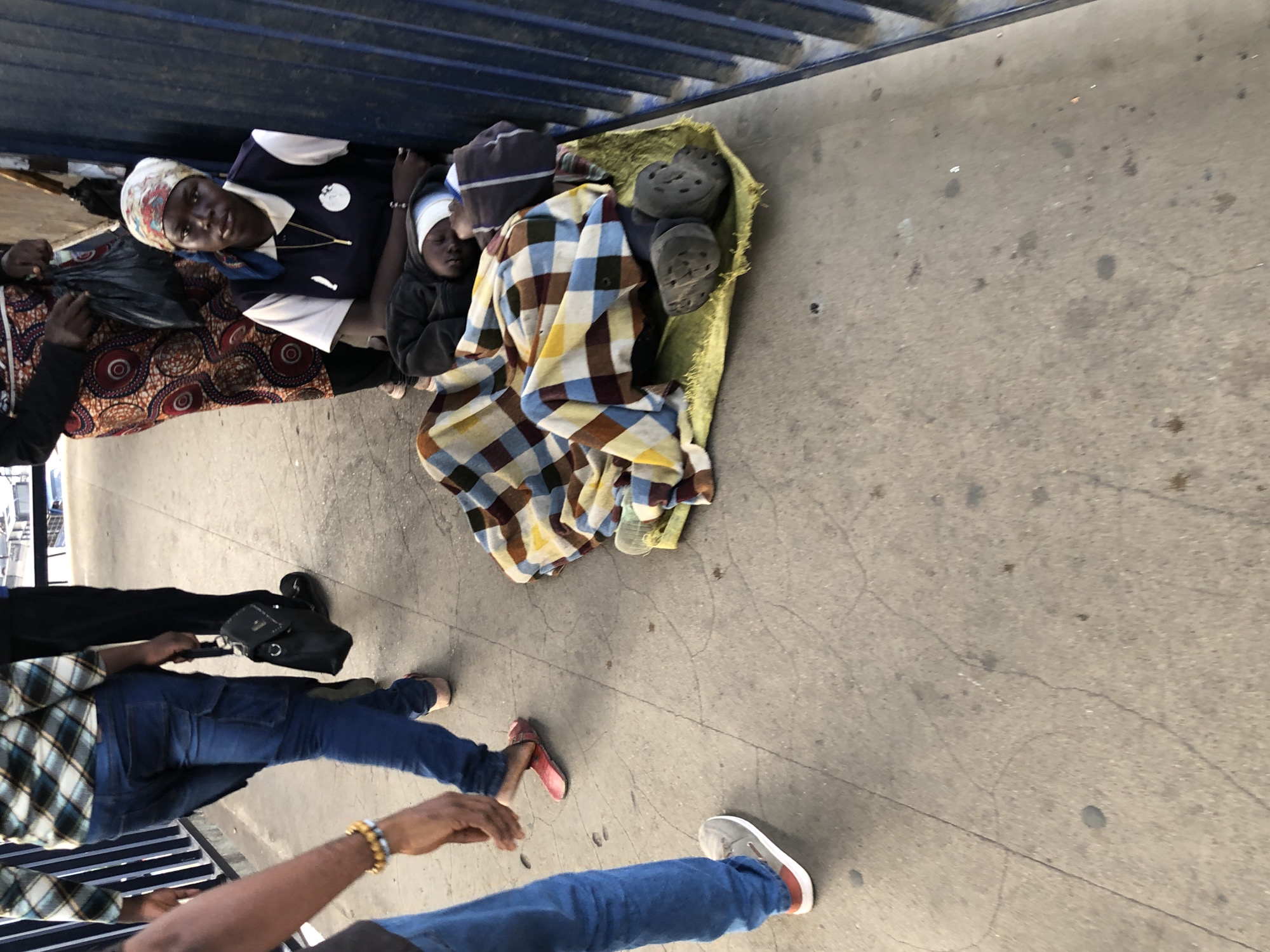 In Ikeja, Nigeria, a street scene unfolds with several individuals present. To the left, a person walks away from the camera, their left leg visible from the knee down, wearing blue jeans and grey sneakers with a red stripe. They are accompanied by another individual to their left, only their arm and hand with a beaded bracelet are visible, also wearing blue jeans. In the center background, a person in a plaid shirt and blue jeans walks away, carrying a black bag. To the right, a woman sits leaning against a blue metal fence, wearing a white and blue collared shirt with a gold chain and a headscarf. She is looking towards the camera. In front of her, another person is lying down, covered by a multi-colored plaid blanket and wearing dark shoes. They are on a yellow mat. The ground is a concrete walkway with visible cracks and some dark stains.