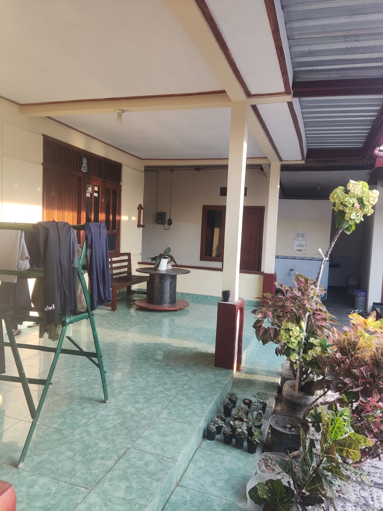 The image depicts an outdoor patio area in Trihanggo, Indonesia. The patio has a tiled floor and a covered roof supported by columns. To the left, a green drying rack holds several pieces of dark clothing. In the background, a wooden bench sits near a wooden door. In the center of the patio, a round wooden table with spools as legs holds a potted plant. To the right, a collection of potted plants, including a tall one with yellow flowers and spiky stems, are arranged along the edge of the patio. The scene is illuminated by natural sunlight.