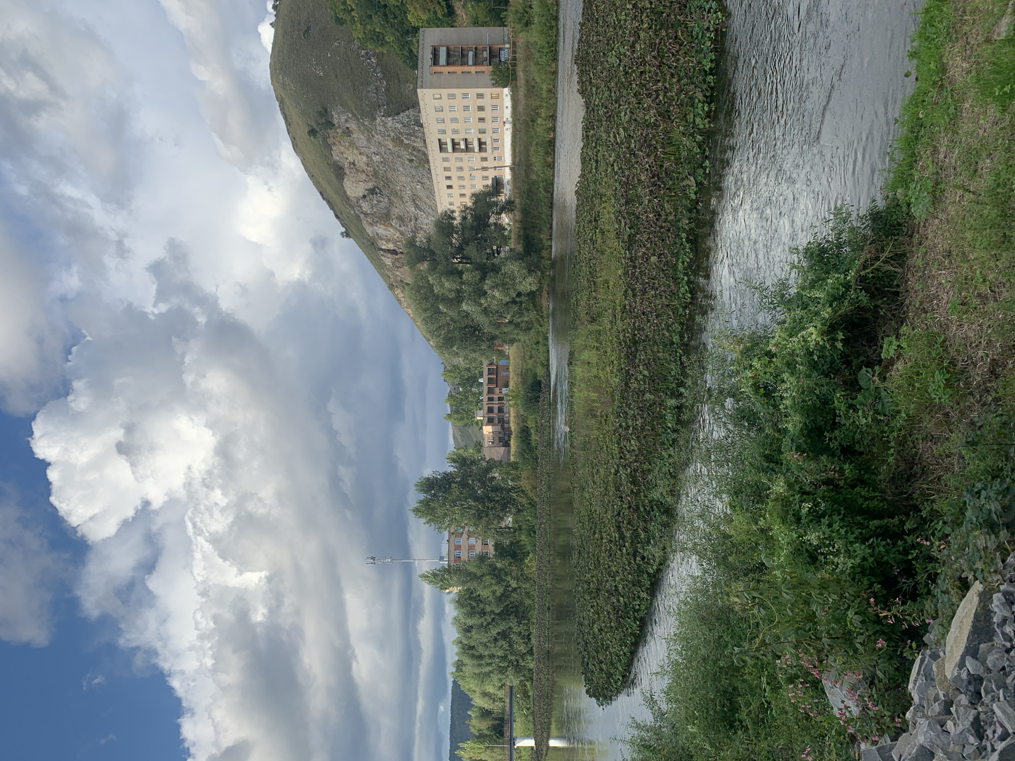 The image depicts a natural landscape with man-made structures in the city of Asha, Russia. In the foreground, a river flows, bordered by lush green vegetation and a pile of grey rocks on the left bank. A prominent feature of the river is a large patch of dense green vegetation growing on its surface, forming a distinct island or mat.
Across the river, a multi-story apartment building with light-colored walls and multiple windows stands against a backdrop of a green, rocky hillside. Another, smaller building with a darker facade is visible to the left of this main building, partially obscured by trees. Further to the left, a slender white tower, possibly a communication mast, rises above a cluster of trees. The sky is filled with dramatic, textured clouds, with patches of blue sky visible in the upper left. The overall scene suggests a somewhat overcast day with diffused sunlight. The environment is a blend of natural elements—river, vegetation, hillside—and urban infrastructure. There are no people visible in the image.