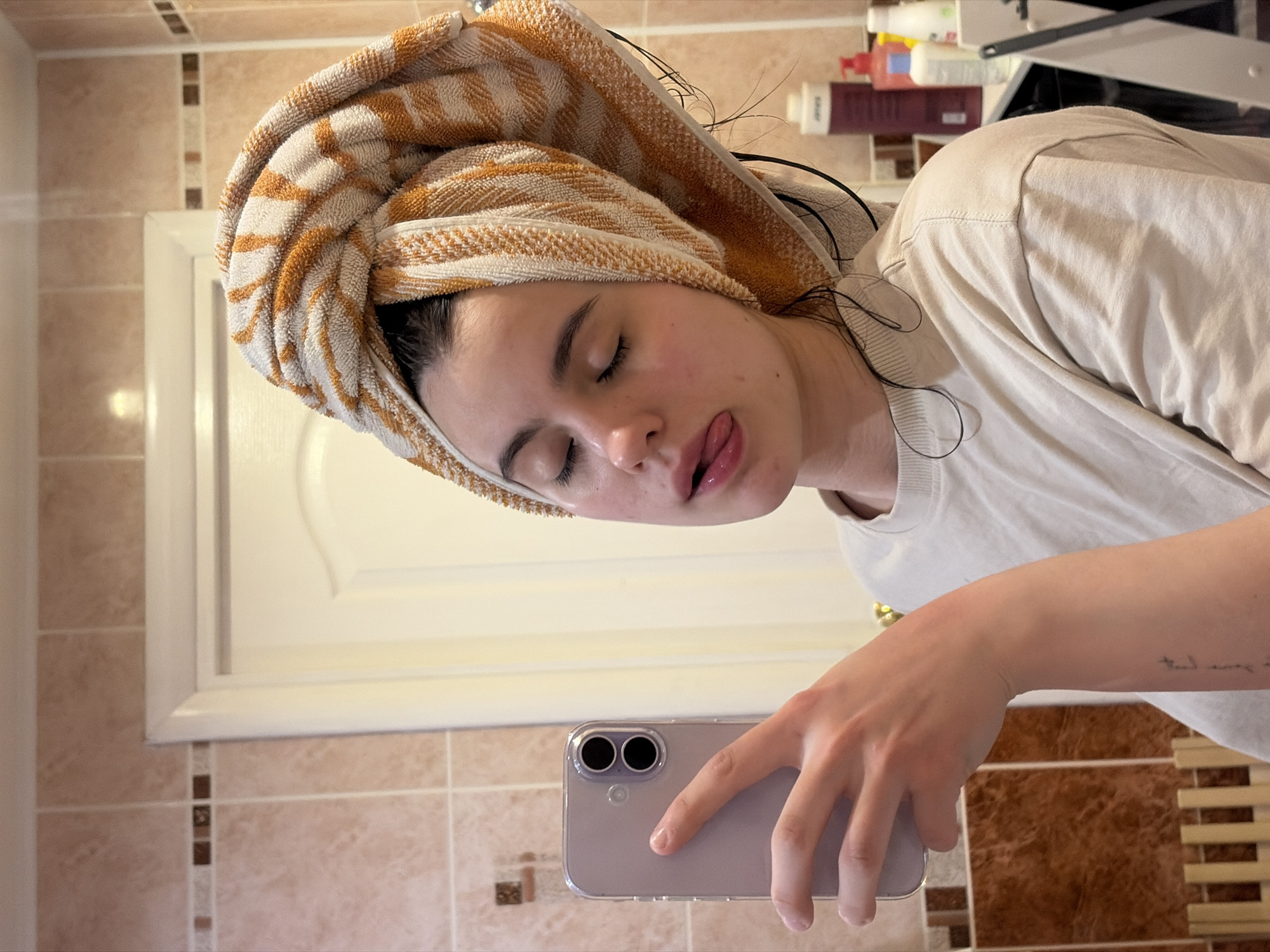 A young woman is taking a selfie in a bathroom. She is wearing a cream-colored t-shirt and has a striped towel wrapped around her head. Her eyes are closed and she is sticking her tongue out. She is holding a light purple phone in her right hand. The bathroom has beige tile with brown accents. There is a white cabinet behind her. The location is Chaikovsky, Russia.