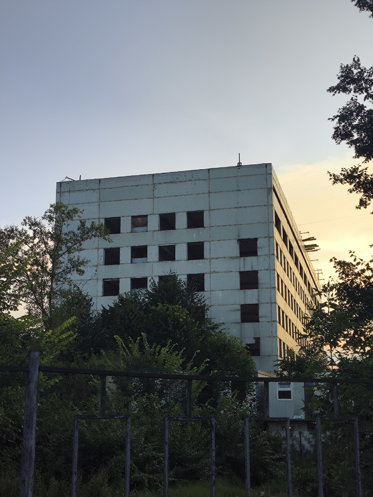 An exterior, eye-level, full shot captures an imposing, multi-story, concrete apartment building against a light blue sky with a hint of yellow sunset on the right. The building appears unfinished or abandoned, with numerous empty, dark window frames dotting its facade. Trees with dark green foliage partially obscure the lower sections of the building, and a rusted metal fence runs across the foreground. A small, portable cabin with a single window is visible behind the fence and in front of the main building. The overall scene conveys a sense of desolation and neglect.