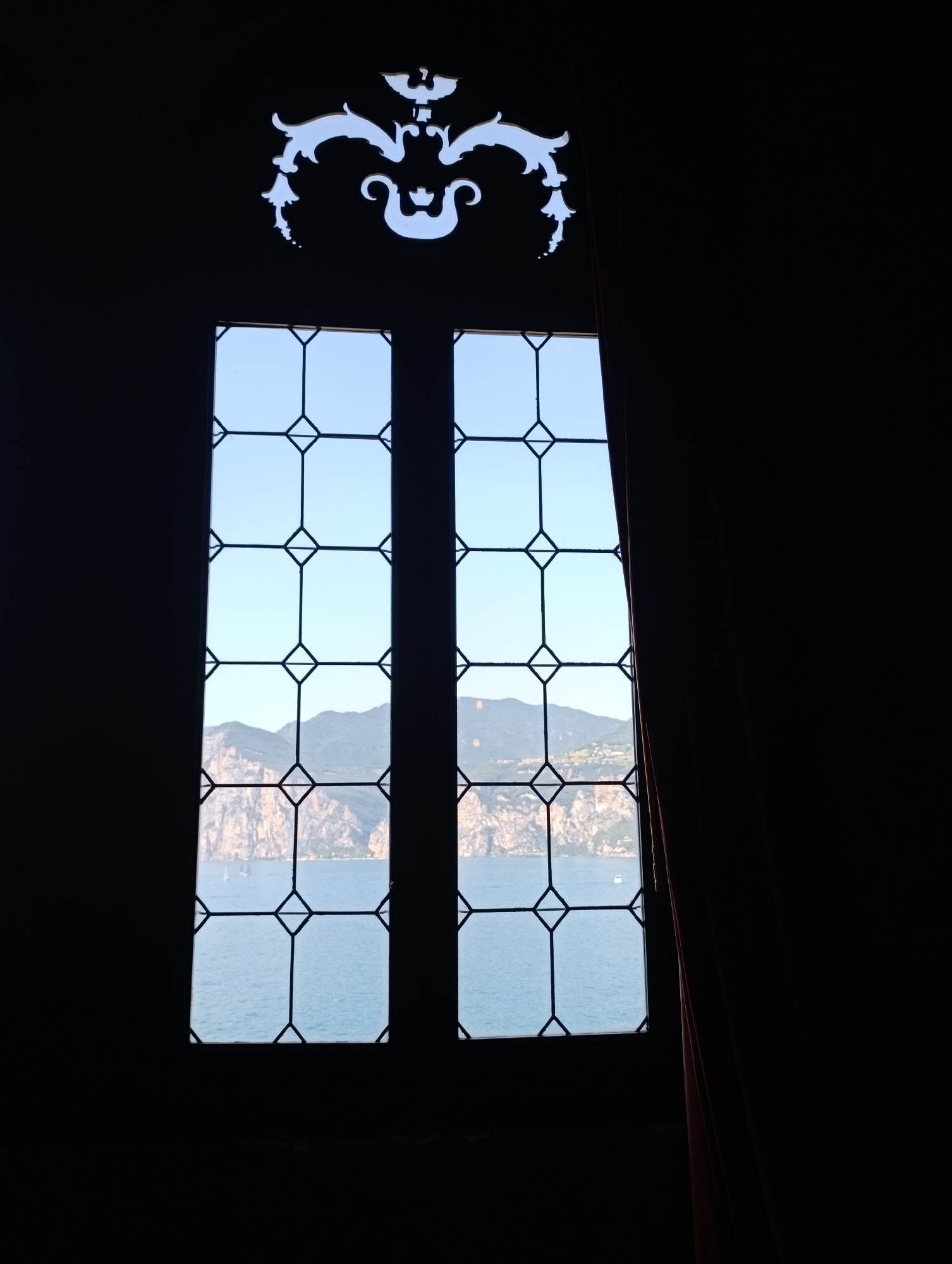 This image, taken from inside a building, looks out through a tall, narrow window with a decorative leaded glass design at the top. The window is divided into two vertical panes, each containing a grid of diamond-shaped panes. Through the window, a body of water, likely a lake, is visible, with steep, rocky mountains rising from its shores. Several sailboats dot the water. A dark red curtain is visible to the right of the window, obscuring a portion of the view. The ornate decoration above the window suggests the building may be historic or elaborately designed. While the specific location in Scandiano, Italy, is not evident from the image itself, the scene evokes a picturesque, possibly lakeside, setting.