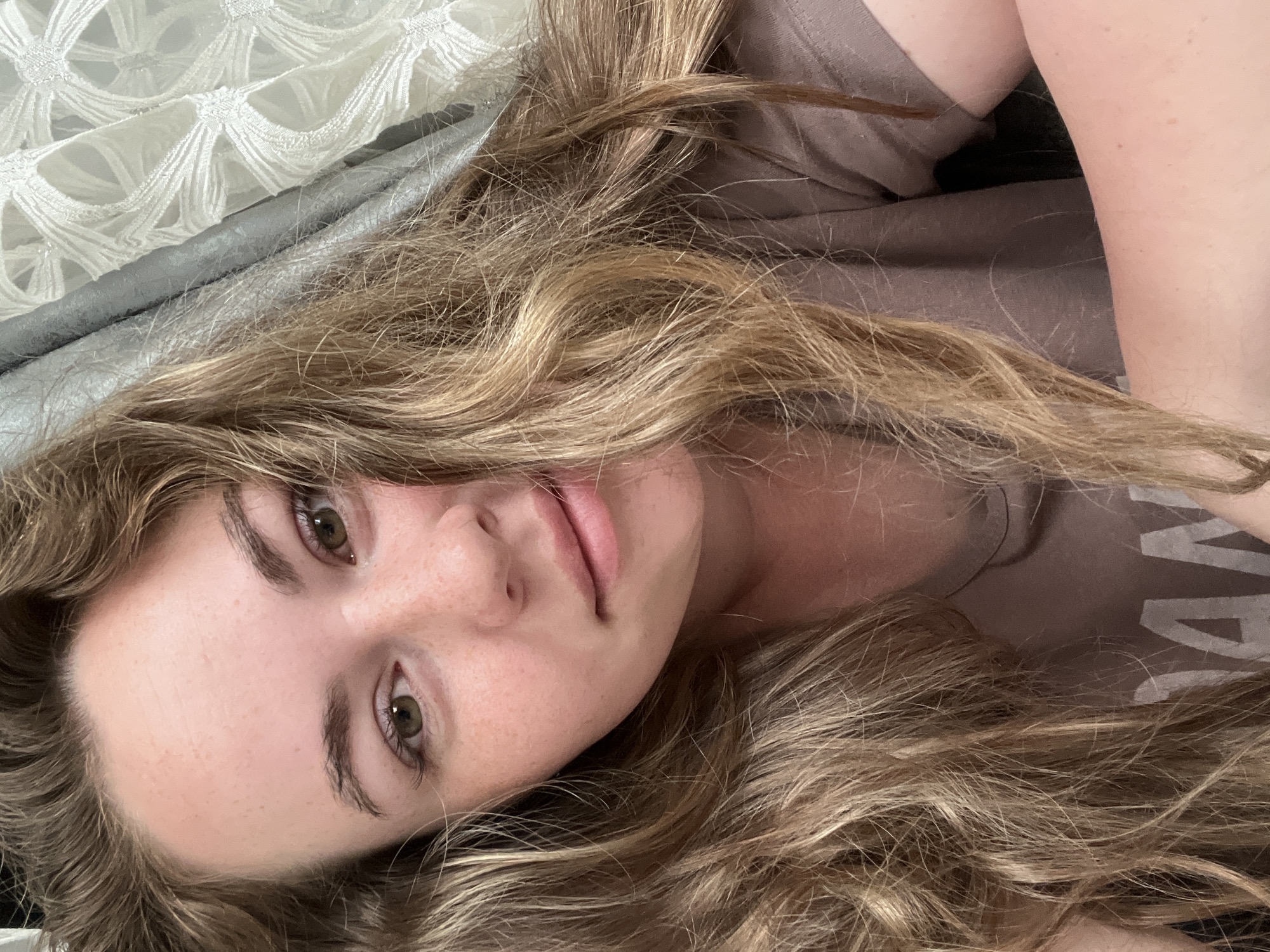 A fair-skinned woman with long, wavy blonde hair is lying on a bed. Her head is turned slightly towards the camera. She has green eyes, freckles, and light pink lips. She is wearing a brown t-shirt. The bed has a white patterned bedspread and a silver-gray sheet underneath.
The location in the city of Chaikovsky, Russia is unknown.