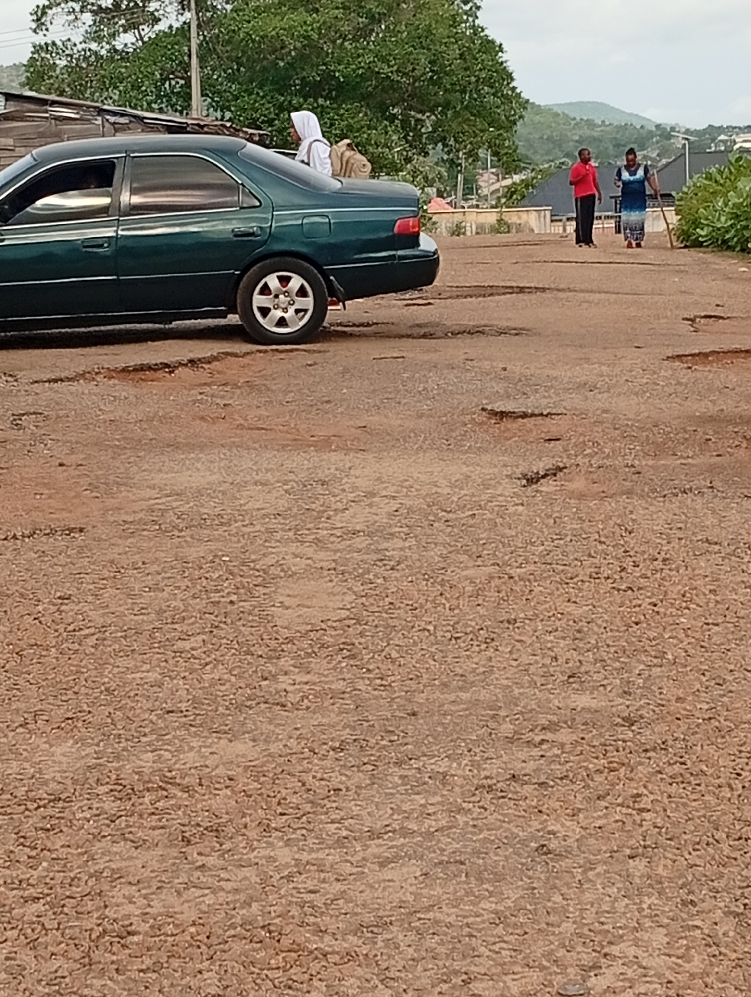 The image depicts a dark green sedan on a badly potholed road in Adankolo, Nigeria. A woman in a white hijab and a backpack is near the rear of the car. In the background, a man in a red shirt and a woman in a patterned blue dress are walking away from the camera. Houses and trees are visible in the background, along with a distant mountain. The road surface is heavily damaged with numerous potholes of varying sizes. The sky is overcast.