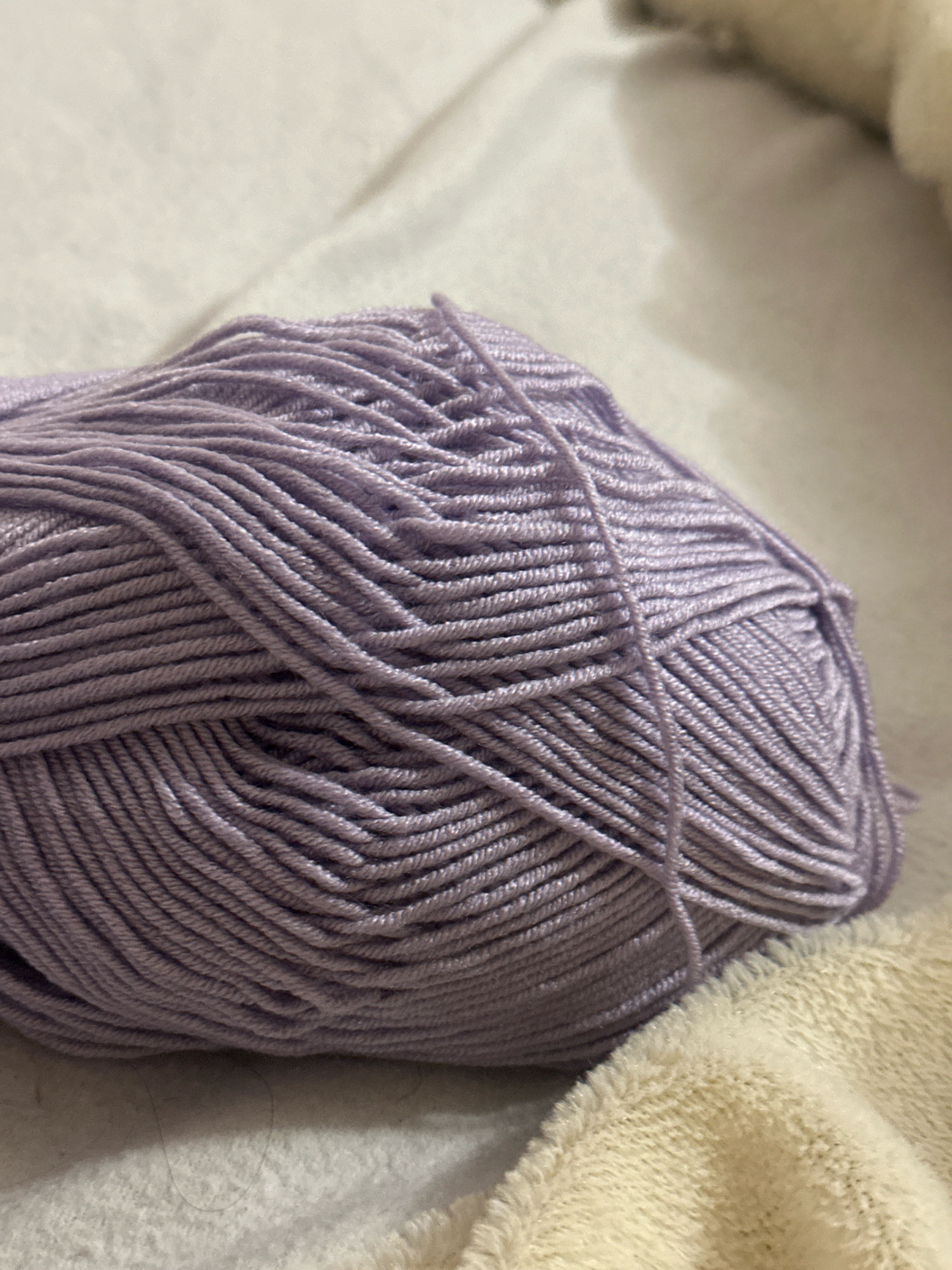 The image shows a close-up of a ball of light purple yarn. The yarn is tightly wound, creating a textured surface with visible strands. A single strand of yarn is loose and trails off to the right. The yarn is placed on a light-colored, textured surface that appears to be fabric, possibly a blanket or towel, which is creamy or beige in color and has a soft, fluffy texture. The background is a light, neutral color and out of focus, suggesting the image was taken indoors with shallow depth of field. There are no people or specific landmarks visible in the image, therefore it is not possible to determine its location in Gyumri, Armenia.