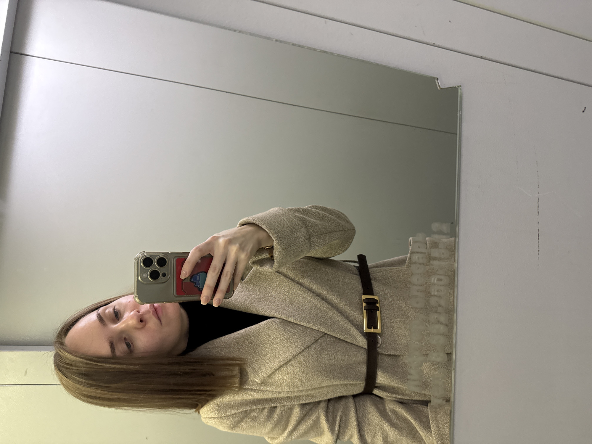 A woman is taking a selfie in a bathroom mirror. She is wearing a beige coat with a dark brown belt. Her hair is light brown and shoulder-length. The bathroom has white walls and a white ceiling with seams visible. The location is Perm, Russia.