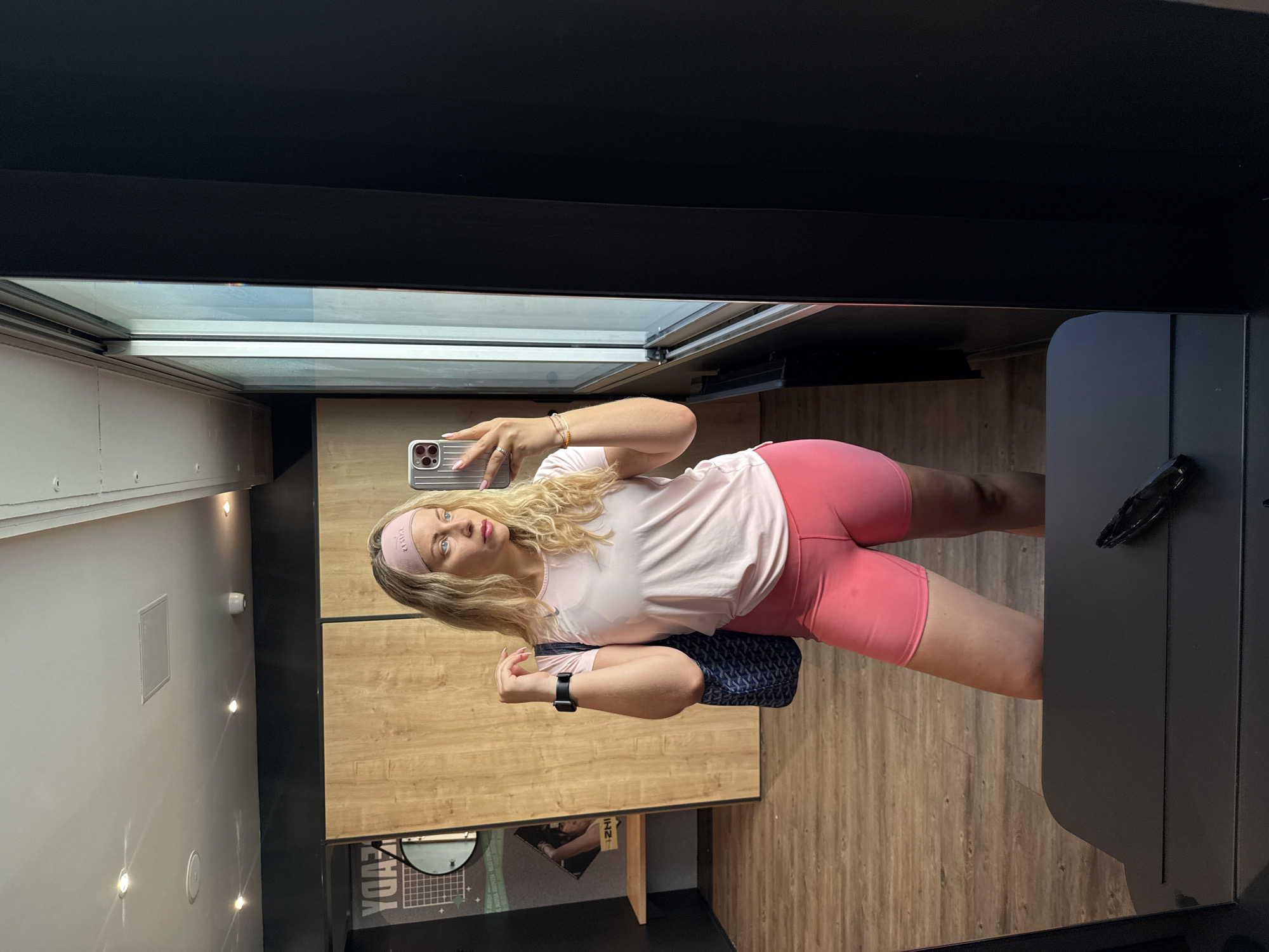 A fair-skinned woman with blonde, wavy hair is taking a mirror selfie in a changing room. She is wearing a pink headband, a light pink t-shirt, and matching pink biker shorts. She is holding a smartphone in her right hand. A dark blue patterned shoulder bag is draped over her right shoulder. The changing room has wooden paneling on one wall and a large mirror on the adjacent wall. The floor has a wood grain pattern. Based on the "Gymshark" branded objects, the image was likely taken in the Gymshark store in Paris, France.