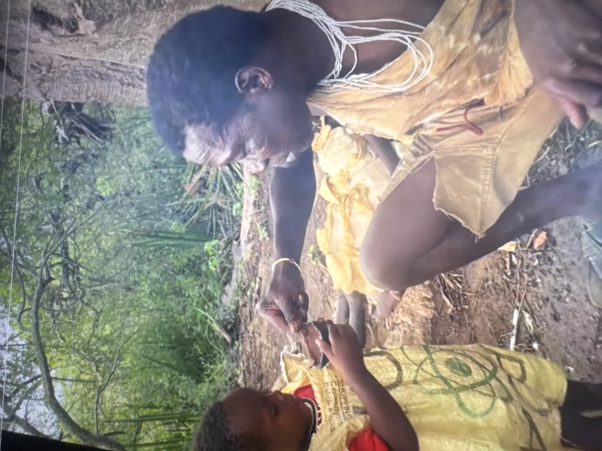 The image depicts two individuals, an adult and a child, engaged in an activity outdoors, possibly in a natural, wooded area. The adult, a male with dark skin and a shaved head, is wearing a necklace made of white beads and a garment that appears to be made of rough, yellowish fabric, possibly animal hide, draped over his shoulders and body. He is seated, facing towards the child. The child, who has dark skin and is looking down, is wearing a yellow garment with a red undershirt. The yellow garment has a pattern of green lines and circles, one of which resembles an atom. The child is holding a small, dark brown, cup-like object in their left hand. The adult's right hand is extended towards the child, holding a small amount of a dark substance, possibly food or medicine, which they appear to be offering to the child. The background suggests a natural environment with trees, foliage, and a dirt ground. There is no visible indication that this scene is located in Abuja, Nigeria, nor are there any urban elements present.