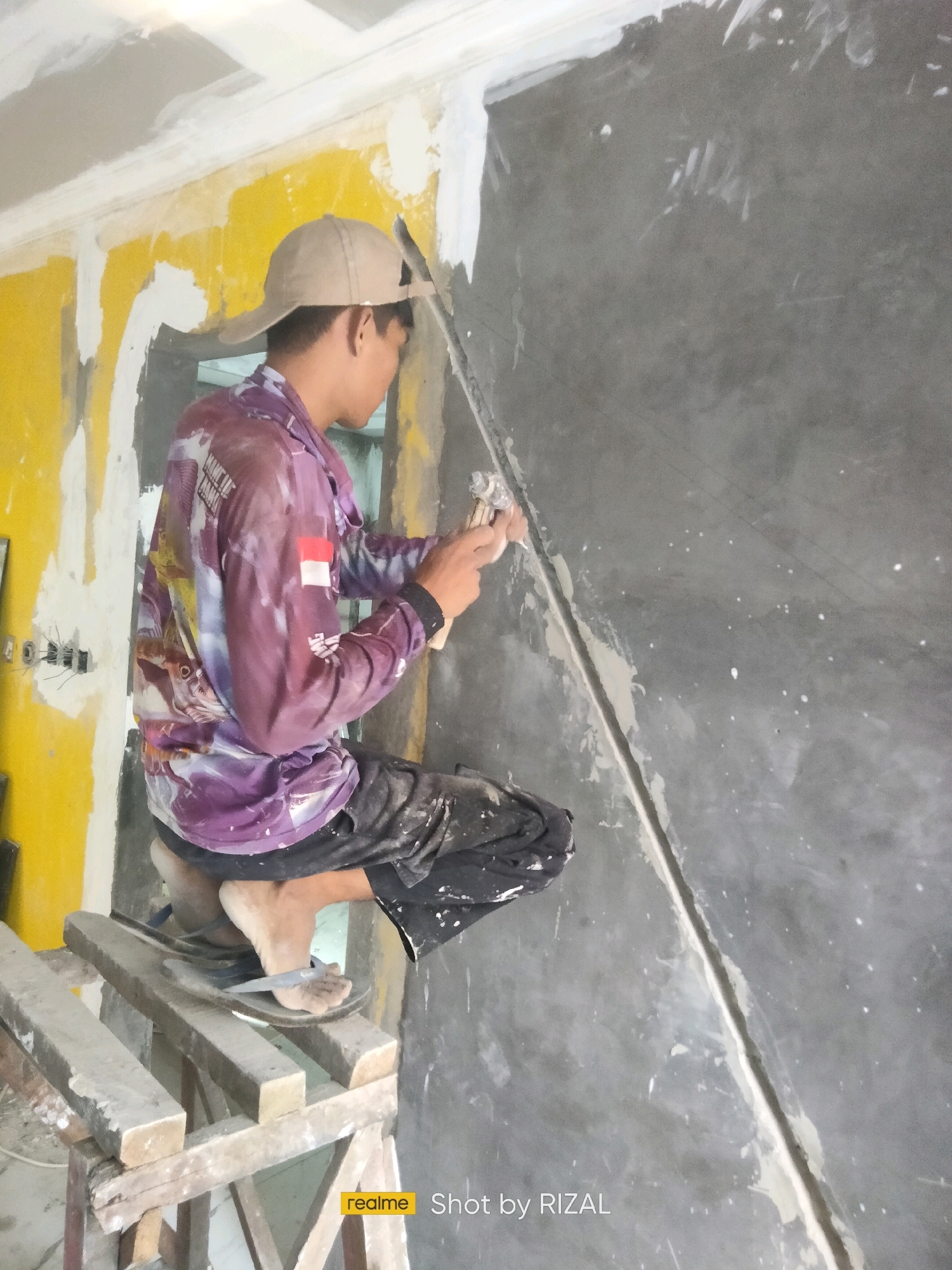 A person wearing a light brown baseball cap and a long-sleeved shirt with a colorful fish print and the Indonesian flag on the sleeve kneels on a wooden support. They are applying a plaster-like substance to a wall joint with a trowel. The wall is dark gray, and there are white speckles on its surface. The background wall is yellow and white, with exposed electrical conduits. The person is wearing dark shorts and sandals. The scene appears to be an indoor construction or renovation site.