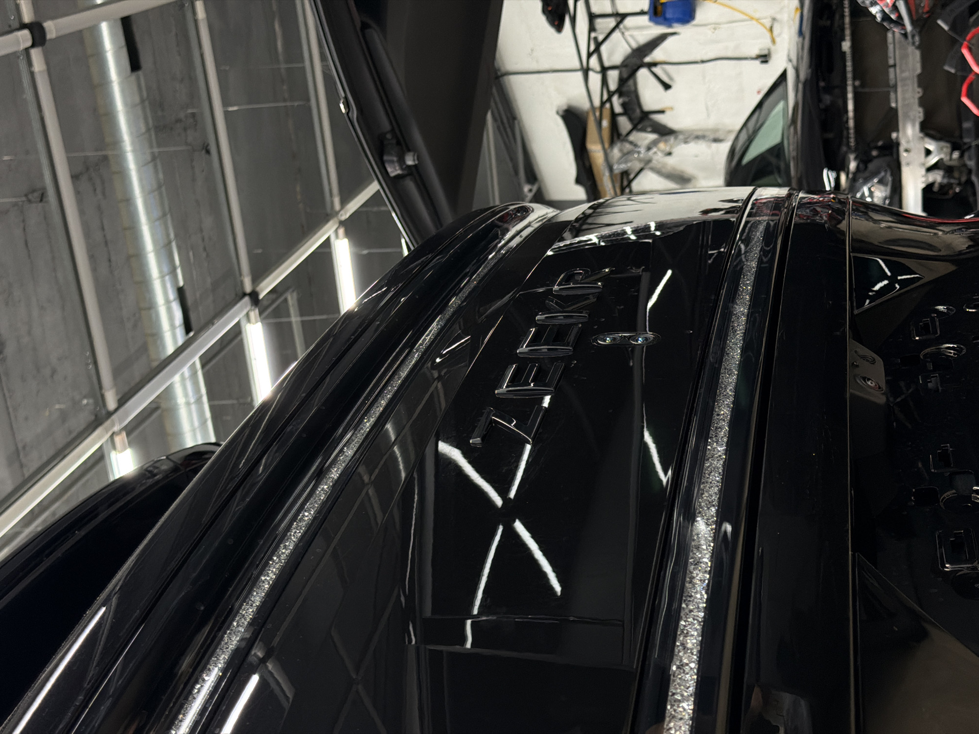 The image presents a detailed, high-angle view of the rear upper section of a black vehicle, likely a luxury or sports car, within an indoor setting in Krasnoyarsk, Russia. No people are visible.
The primary subject is the black car, which features an extremely glossy, polished finish that reflects the surroundings vividly. Visible on its surface are the metallic letters "ETE," part of an emblem or branding. Two prominent strips with a textured, sparkling, or 'diamond-like' appearance run along channels on the car's body, catching the light brightly. The highly reflective surface of the vehicle also reflects overhead fluorescent lights, creating distinct "X" shapes and linear reflections.
The setting appears to be a garage, workshop, or detailing studio. The background reveals an industrial-style ceiling with visible concrete, metal framework, long fluorescent light tubes, and a large, round, silver ventilation duct. In the upper right, a cluttered area with wires, equipment, and possibly other car parts is visible, along with a partially obscured vehicle in the blurred distance.
The car is stationary, and no specific activity is occurring. The strong artificial lighting emphasizes the vehicle's pristine condition and the intricate details of its design, suggesting it might be prepared for display or recently detailed.