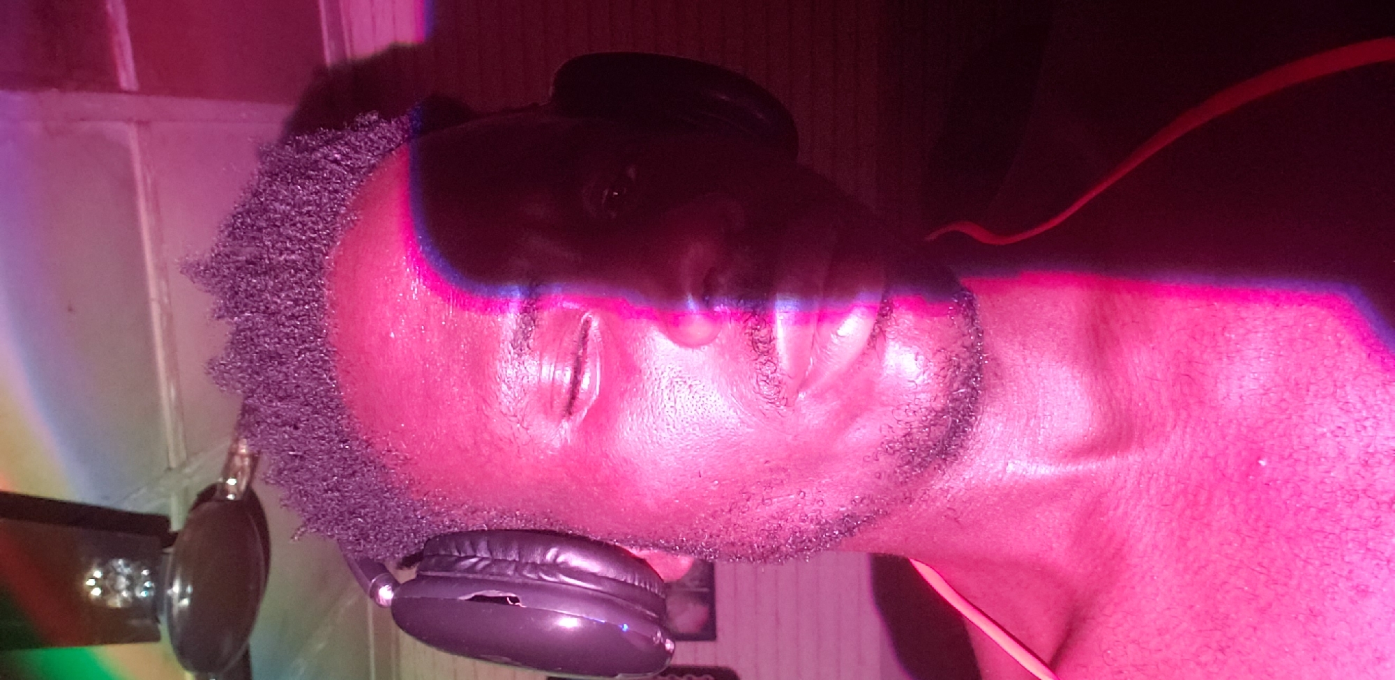 The image is a close-up, waist-up shot of a Black man wearing headphones, taken from a low angle. The man has short, dark, curly hair and a light beard. His left eye is closed, and his right eye is partially open. He is wearing a thin, light-colored strap across his chest.
The scene is illuminated by vibrant pink and purple lighting, with a distinct vertical stripe of bright pink light bisecting the man's face from his forehead to his chin. A dark, shadowed area covers the right side of his face. In the upper left corner of the image, a portion of a ceiling fan is visible, with its blades casting faint, colorful reflections. The background appears to be a wall, with some texture visible, possibly brickwork. A faint image of what looks like a framed picture can be seen on the wall to the left of the man's head.
The overall impression is that of a person enjoying music or a party atmosphere, given the lighting and the headphones. The photograph seems to have been taken indoors. The location in Narayi, Nigeria is not directly depicted in the image itself, but is provided as context for the scene.