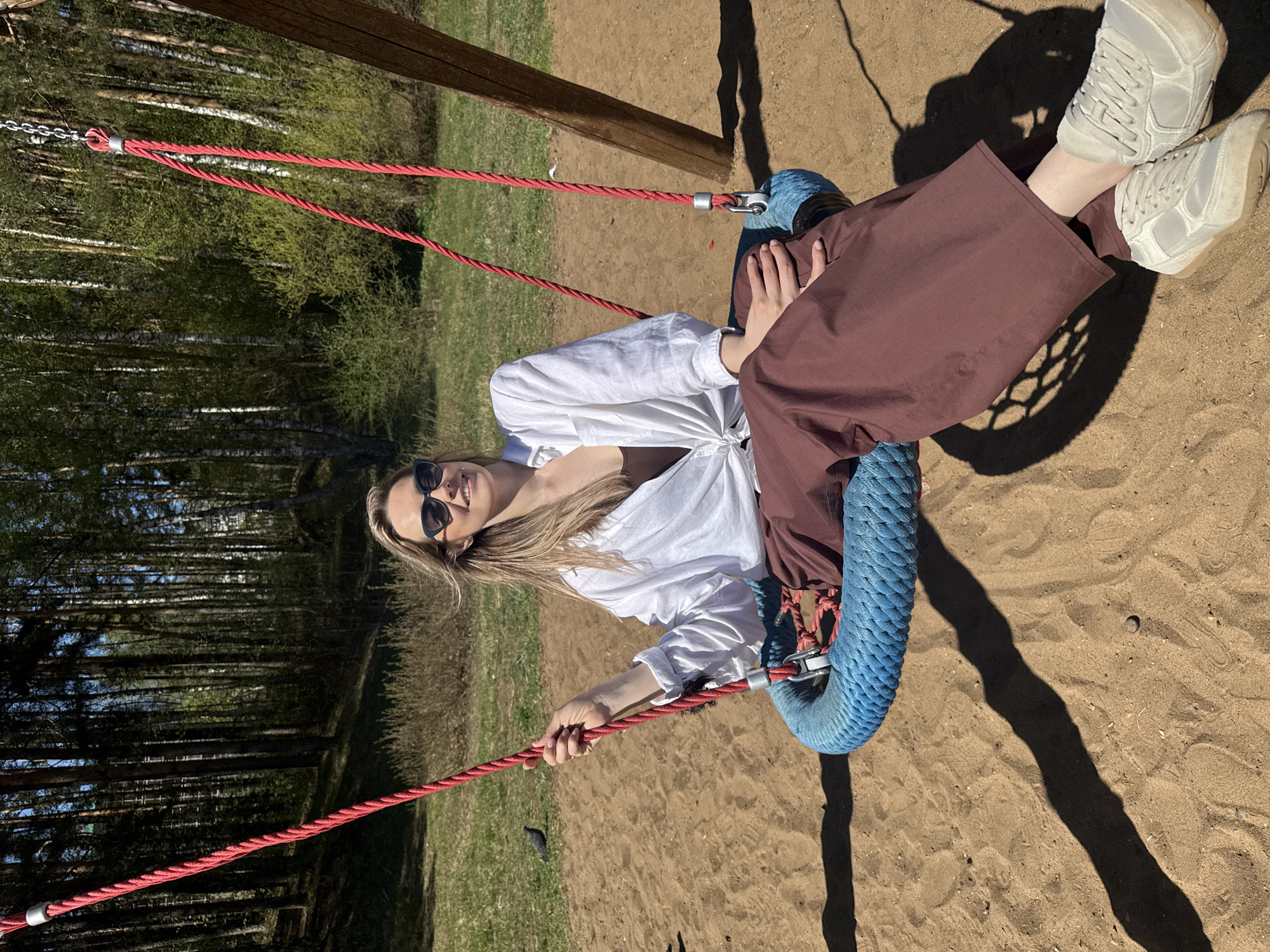 A woman sits in a blue tire swing in Chaikovsky, Russia. She wears a white shirt, brown pants, and white sneakers. The swing is suspended by red ropes attached to a wooden frame. The ground is sandy, and trees are visible in the background.