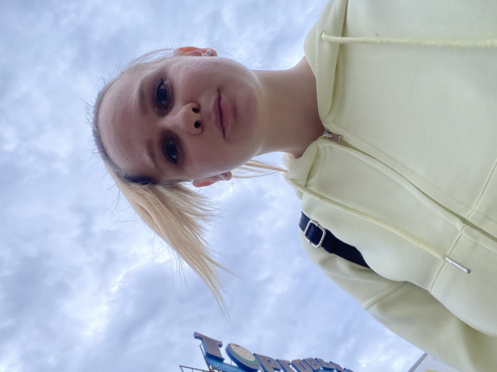 A low-angle, upward-facing shot captures a young woman with light skin and fair hair pulled back in a low ponytail. She's wearing a pale yellow hooded sweatshirt with a black strap visible over her shoulder. Behind her, the sky is overcast with gray clouds. A building sign, partially obscured by the angle, is visible at the bottom of the frame, with the letters "TOP" discernible in blue. The sign's structure and style suggest an amusement park or similar recreational facility. Given the location of Chaikovsky, Russia, it is likely that the amusement park is close by.