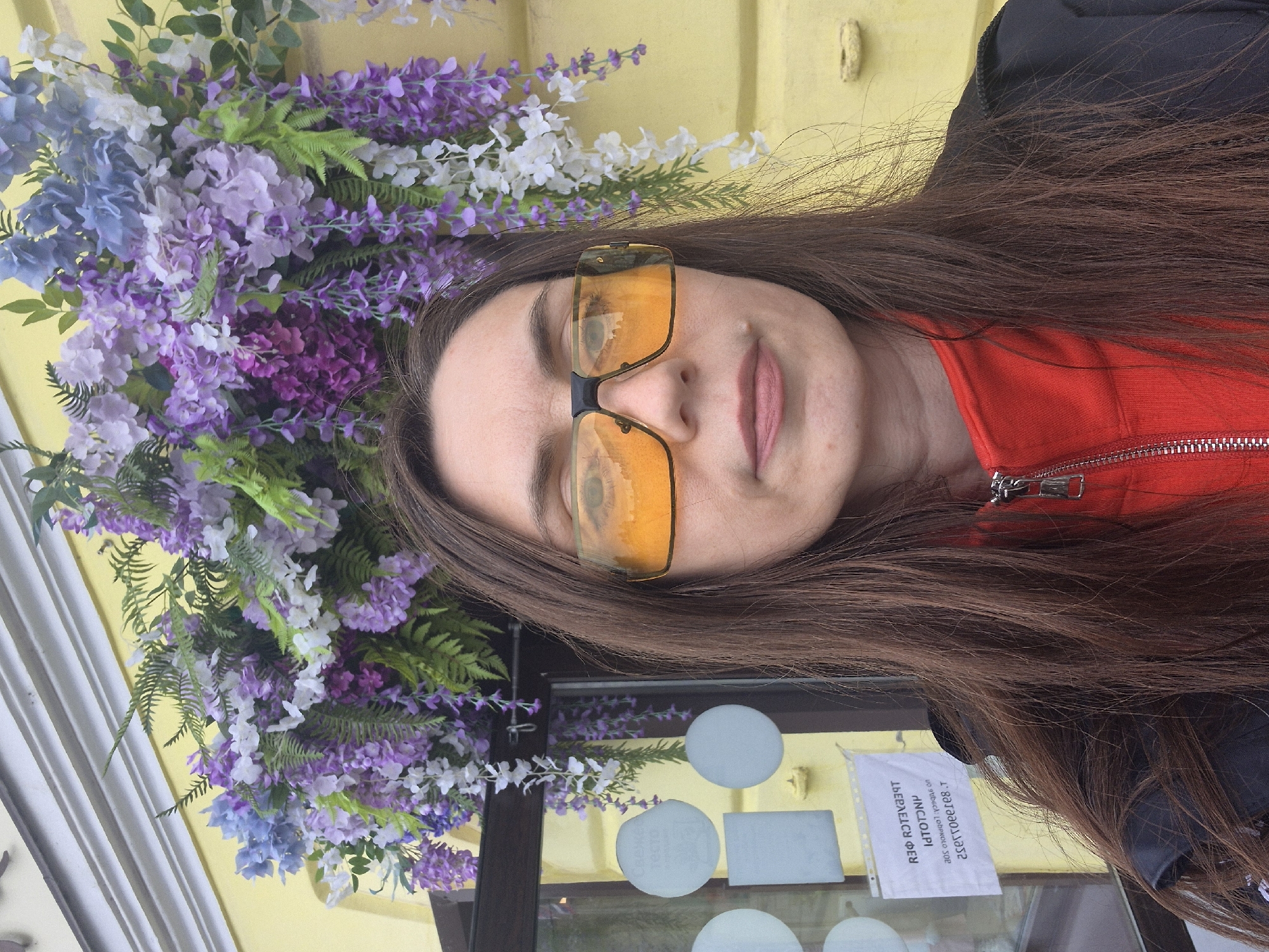 A woman is captured in a close-up, eye-level selfie-style photograph, standing outdoors in Sarapul, Russia. She has long, straight, dark brown hair cascading over her shoulders and wears large, rectangular, orange-tinted sunglasses with a black frame and bridge; her eyes are faintly visible behind the reflective lenses. Her attire consists of a red, collared, zippered top under a black outer garment, visible on her right shoulder (viewer's left). A small, light-colored blemish or mole is visible on her upper lip, below her right nostril (viewer's left).
The background features a light yellow building with white architectural molding along its top edge. An elaborate artificial floral arrangement, dominated by shades of purple, blue, and white wisteria-like and hydrangea-like flowers, interspersed with green fern foliage, hangs above and behind her head, attached to the building. To her left, a dark-framed glass window or door reveals several reflective circular and rectangular white stickers or signs on its surface. One rectangular sign displays inverted Cyrillic text, indicating a Russian-speaking location and confirming the city of Sarapul, Russia, as the specified location.