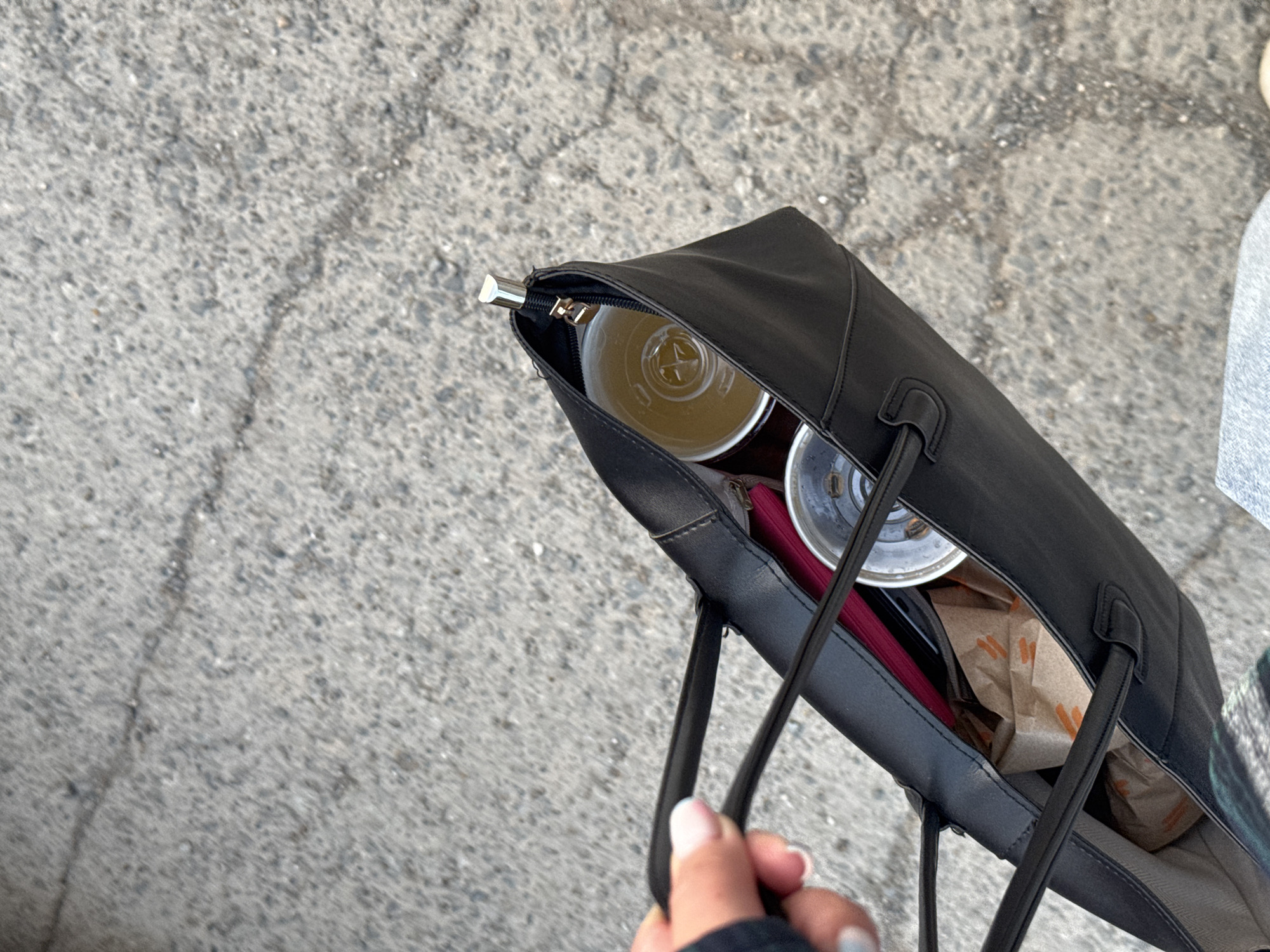 The image shows a top-down view of a black tote bag filled with various items, including two disposable cups with lids, one of which contains a light-colored beverage. There is also a pink object that appears to be a wallet or small pouch, and two crumpled paper bags with orange branding. A person's hand with manicured nails is holding one of the bag's straps. The bag is positioned over a grey, asphalt-like surface, likely a sidewalk or road. The image does not provide any specific details that would allow for the identification of the city of Perm, Russia.