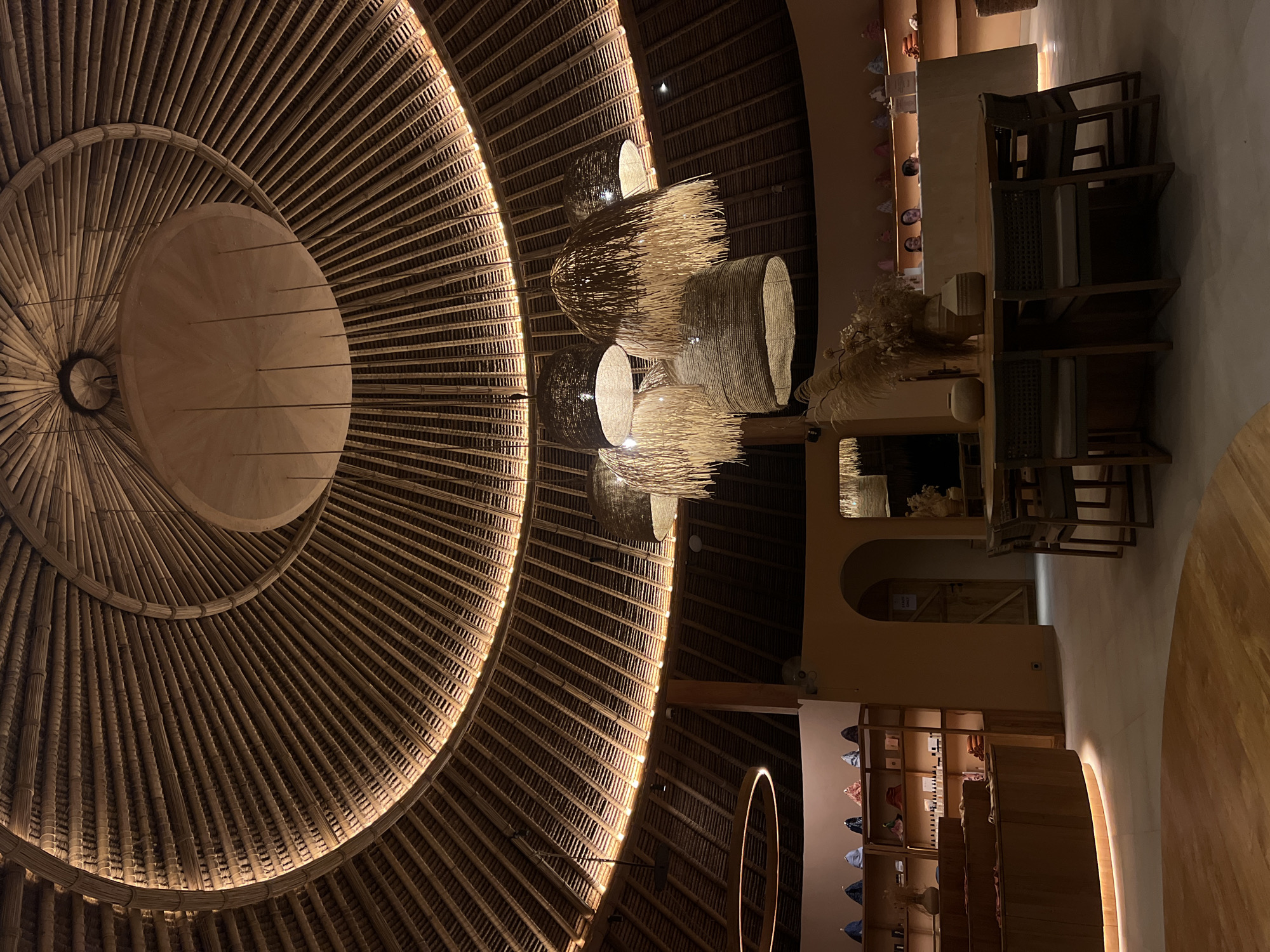 This is an interior shot of a circular, dome-shaped building with a distinctive thatched roof. The roof is constructed of many thin, parallel slats of a natural material, possibly bamboo or straw, radiating from a central point and creating a visually striking ribbed pattern. Integrated lighting runs along the perimeter of the dome, casting a warm glow.
Several large, woven pendant lamps hang from the ceiling, their intricate textures and natural materials complementing the roof's design. Below, the space appears to be a retail or hospitality establishment. In the foreground, a wooden table is set with chairs, suggesting a dining or waiting area. To the left, a curved wooden display counter with integrated lighting is visible, showcasing various items. Behind it, shelves are stocked with merchandise, possibly clothing or accessories, including several hats arranged on top.
Further back, an arched doorway leads to another area, with a wooden door. A reception desk is visible, with at least three individuals behind it, interacting with unseen customers. The walls are painted in a warm, neutral tone. The overall aesthetic is organic, natural, and designed to evoke a sense of calm and possibly a tropical or bohemian ambiance. The image was taken in Kediri, Indonesia, suggesting the architectural style and materials are representative of the region or inspired by its natural elements.