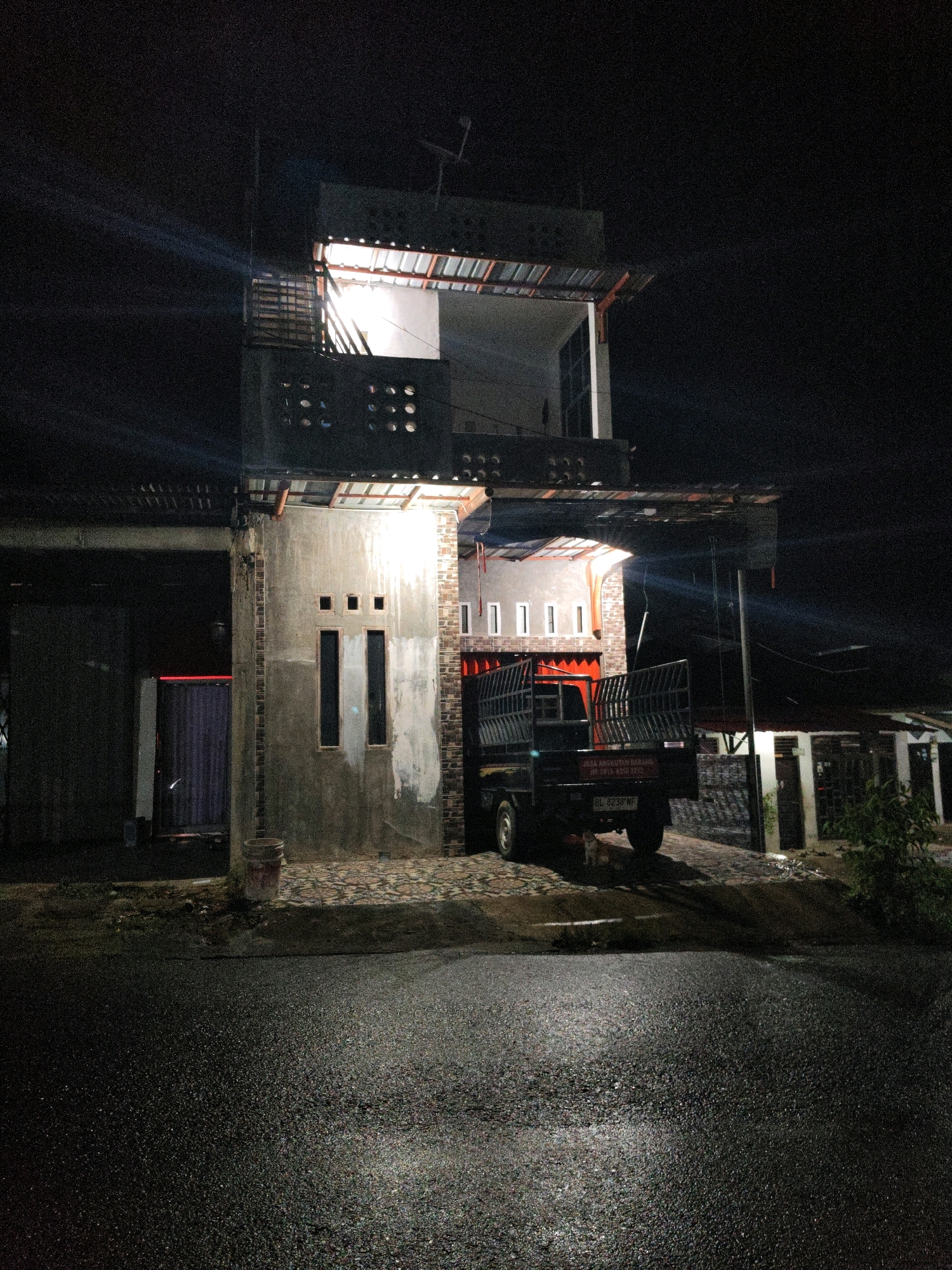 The image depicts a nighttime scene in Lhokseumawe, Indonesia. A two-story building with an unfinished appearance is illuminated by bright artificial lights. The lower level has a covered entryway with a dark colored corrugated metal roof. Adjacent to this is a concrete wall with two tall, narrow windows and three square openings above them. A black pickup truck with a metal cage on the back is parked in front of this structure, partially under an overhang. The truck has license plate number "BL 8238 WF". To the left of the building, there is a dark entryway with vertical blue corrugated metal doors and a red glow emanating from within. The ground in front of the building is paved with a textured material, and further forward, a wet asphalt road reflects the light. In the background, other buildings are visible, also illuminated. A small cat is standing near the rear wheel of the truck. The overall environment appears to be an urban or semi-urban setting at night.