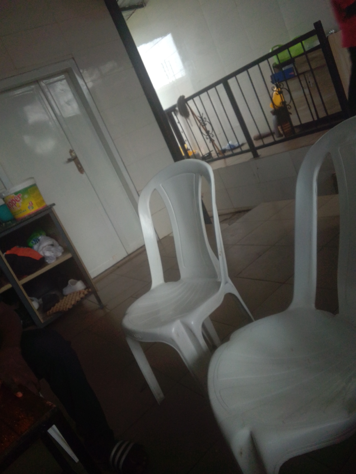 The image shows an indoor scene with two white plastic chairs prominently in the foreground. The chairs are simple, molded designs with curved backs and legs. Behind the chairs, a shelf unit is visible, containing various items including a yellow container with "opper" written on it, some folded clothing, and a pair of white shoes. To the left of the shelf unit is a white door with a gold handle. In the background, a metal railing separates the area from what appears to be a raised platform or balcony. Objects on this platform include a green container and a yellow object, possibly a lamp. The flooring is tiled with a diamond pattern. The lighting suggests it is daytime, possibly indoors with artificial light. The overall impression is of a functional, perhaps utilitarian space.