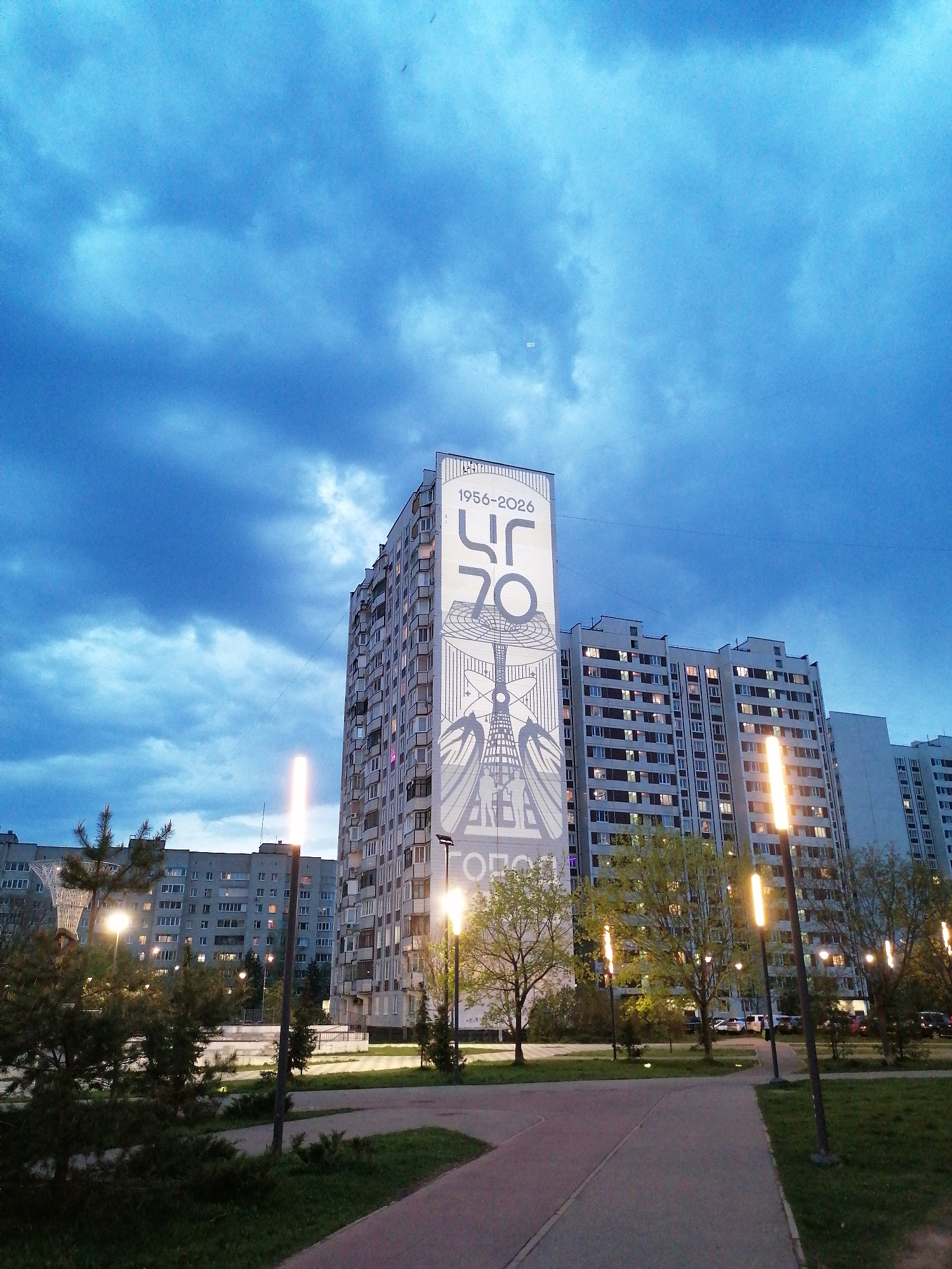 The image shows a street scene at dusk in an urban area, likely in the city of Unknown, Unknown. The sky is a deep blue with dramatic clouds. Several tall apartment buildings dominate the backdrop, with the closest one featuring a large mural on its side, displaying "1956-2026" and "70" alongside other graphic elements. A pedestrian walkway stretches into the distance, flanked by green grass and young trees. Modern street lamps illuminate the walkway with a warm glow. No people are visible in the frame.