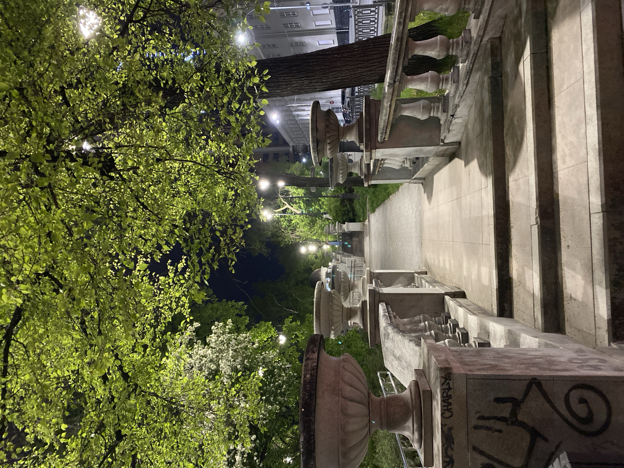 This image shows a pathway at night in Yekaterinburg, Russia. The pathway is lined with ornate stone balustrades and planters. Trees with bright green foliage are visible on the left side of the image, illuminated by streetlights. The pathway leads towards buildings in the distance. Graffiti is present on the stone planters.