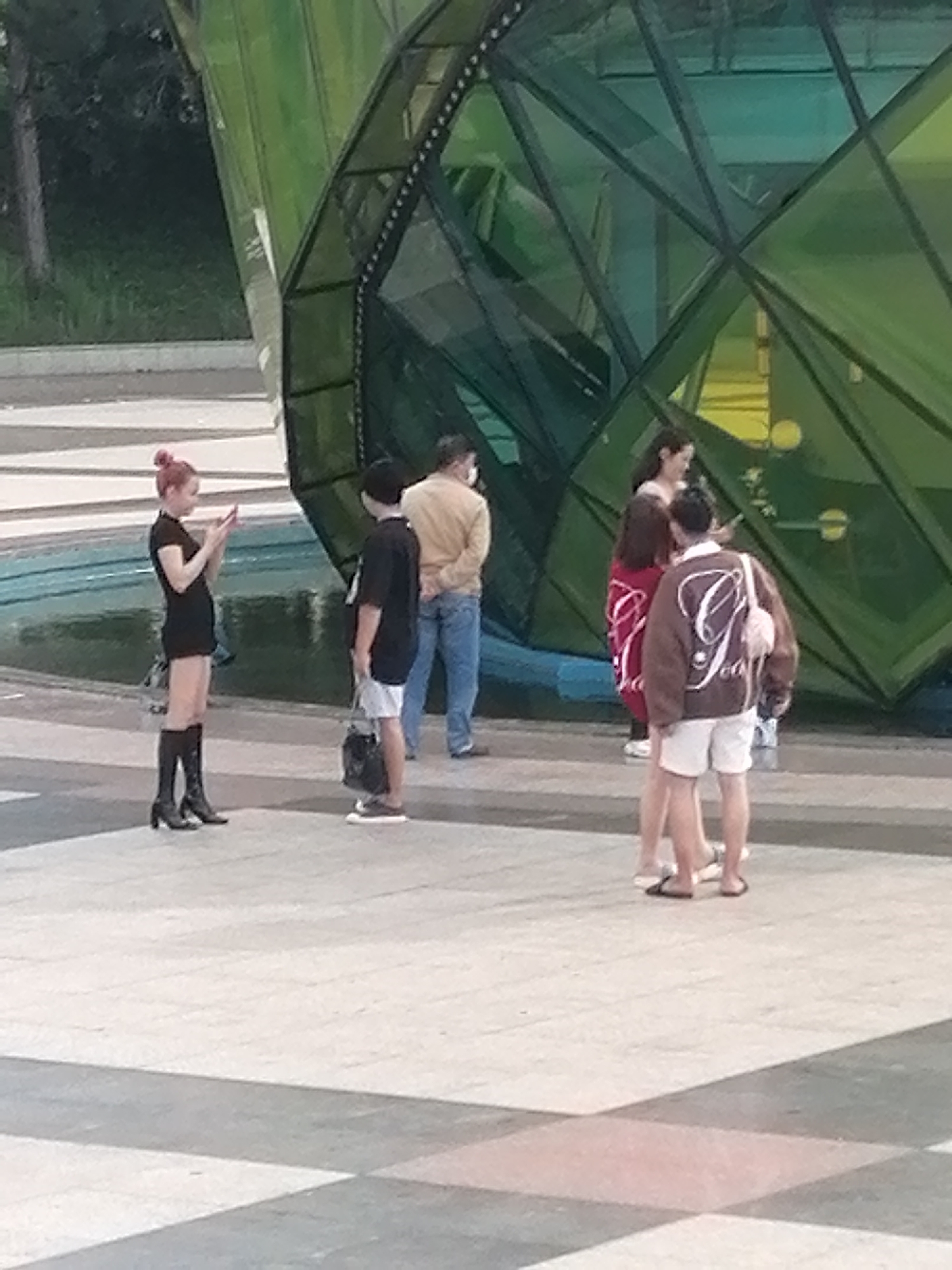 This image depicts a group of people near a distinctive geodesic dome structure with green, faceted panels, likely an architectural feature or part of a public installation. The ground is paved with large, light-colored tiles, some of which are a pinkish-beige hue, creating a patterned effect. In the background, a hint of greenery and trees suggests an outdoor urban setting.
On the left, a person with reddish-pink hair styled in a bun is wearing a short black dress and knee-high black boots. They are holding a smartphone in their hands, appearing to be using it. Beside them, a person in a black t-shirt and light-colored shorts stands facing them, holding a dark bag.
Further to the right, a person in a light brown long-sleeved shirt and blue jeans is visible. Next to them, a person is wearing a dark brown long-sleeved top and a red and white patterned wrap skirt. They appear to be interacting with another person who is wearing a brown jacket with a large white "J" logo on the back and white shorts. This individual is also carrying a small pink bag. The overall impression is a casual gathering in a public space, possibly a park or plaza in Da Lat, Vietnam, given the context of the dome structure and the warm weather attire.