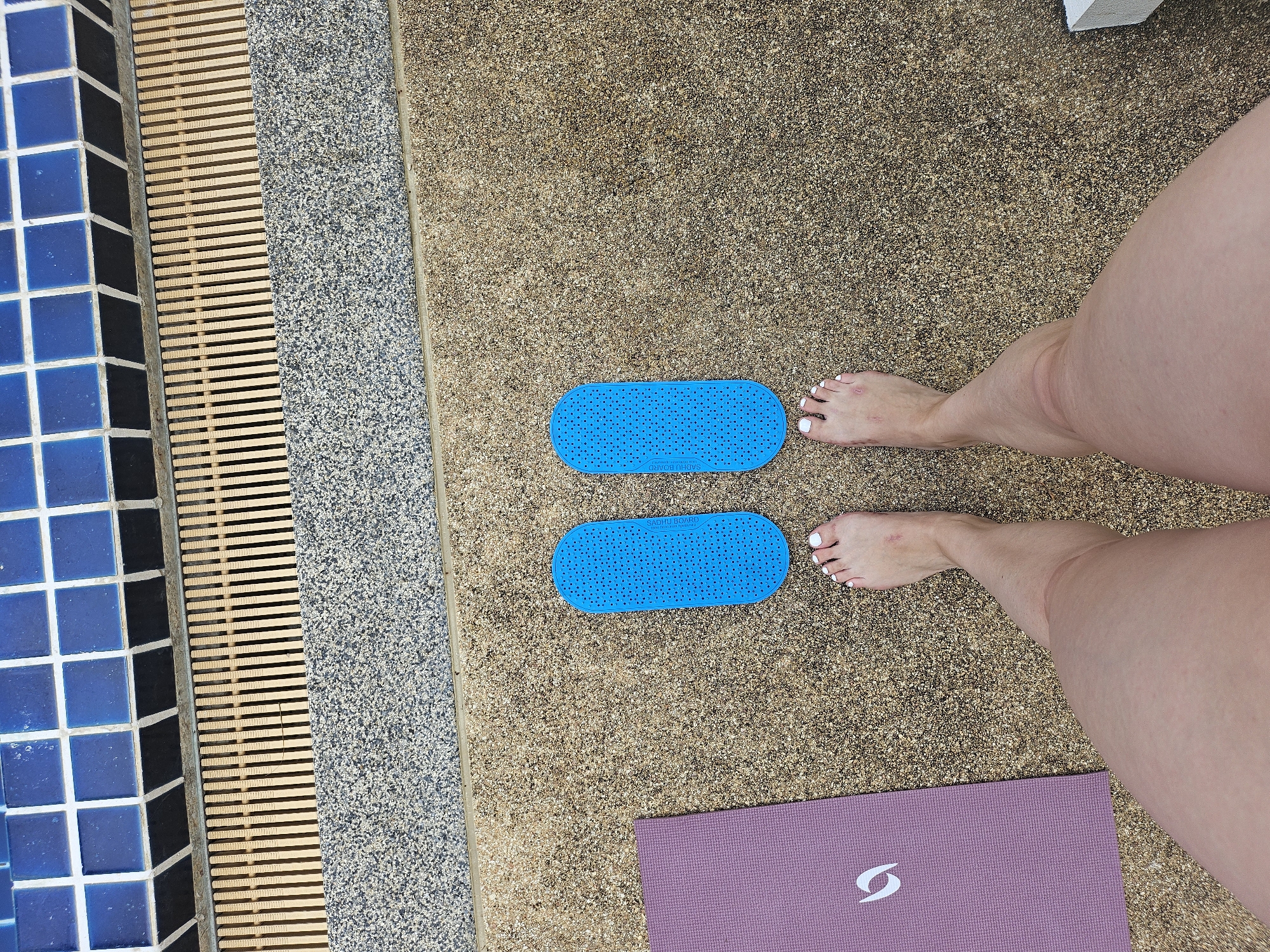 The image depicts a person's feet and legs standing on a textured, pebble-like surface beside a swimming pool. Two oval-shaped blue objects with small holes are positioned in front of the feet. To the left, a portion of a purple yoga mat is visible. The swimming pool edge is lined with blue tiles and a drainage grate. The scene appears to be outdoors, likely in a recreational area. The location is described as Kathu, Thailand.