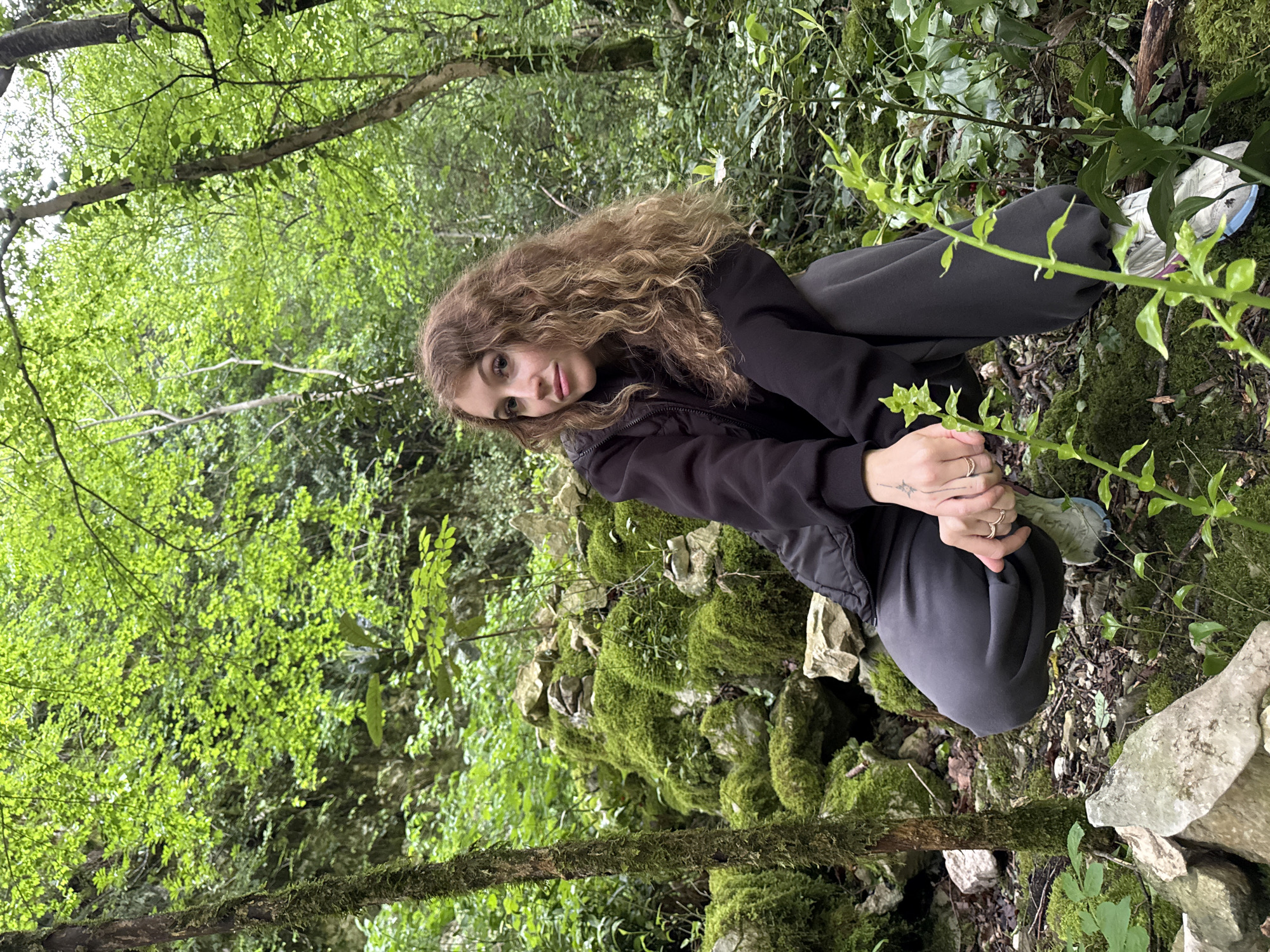 A young woman with long curly hair is crouching in a lush, overgrown area in what could be Tsabal, Abkhazia. She is wearing a dark jacket and dark pants, and she is facing the camera with her hands clasped. There is a moss-covered rock formation behind her and dense greenery all around, including trees and vines. Her location is unidentifiable, as it is not visible in the image. Her facial features are visible, and she has rings on her fingers.