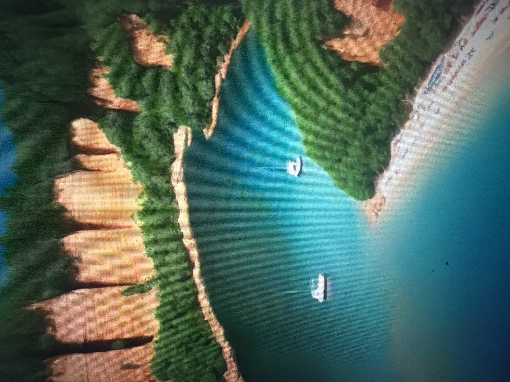 The image depicts a scenic view of a tropical cove with lush green foliage and dramatic orange cliffs surrounding a turquoise body of water. Two white sailboats are visible in the water, one in the foreground and another further in the distance. A sandy beach lines the edge of the cove, dotted with what appear to be beach chairs or loungers. The overall impression is one of a serene and picturesque coastal landscape, characteristic of popular tourist destinations in Thailand.