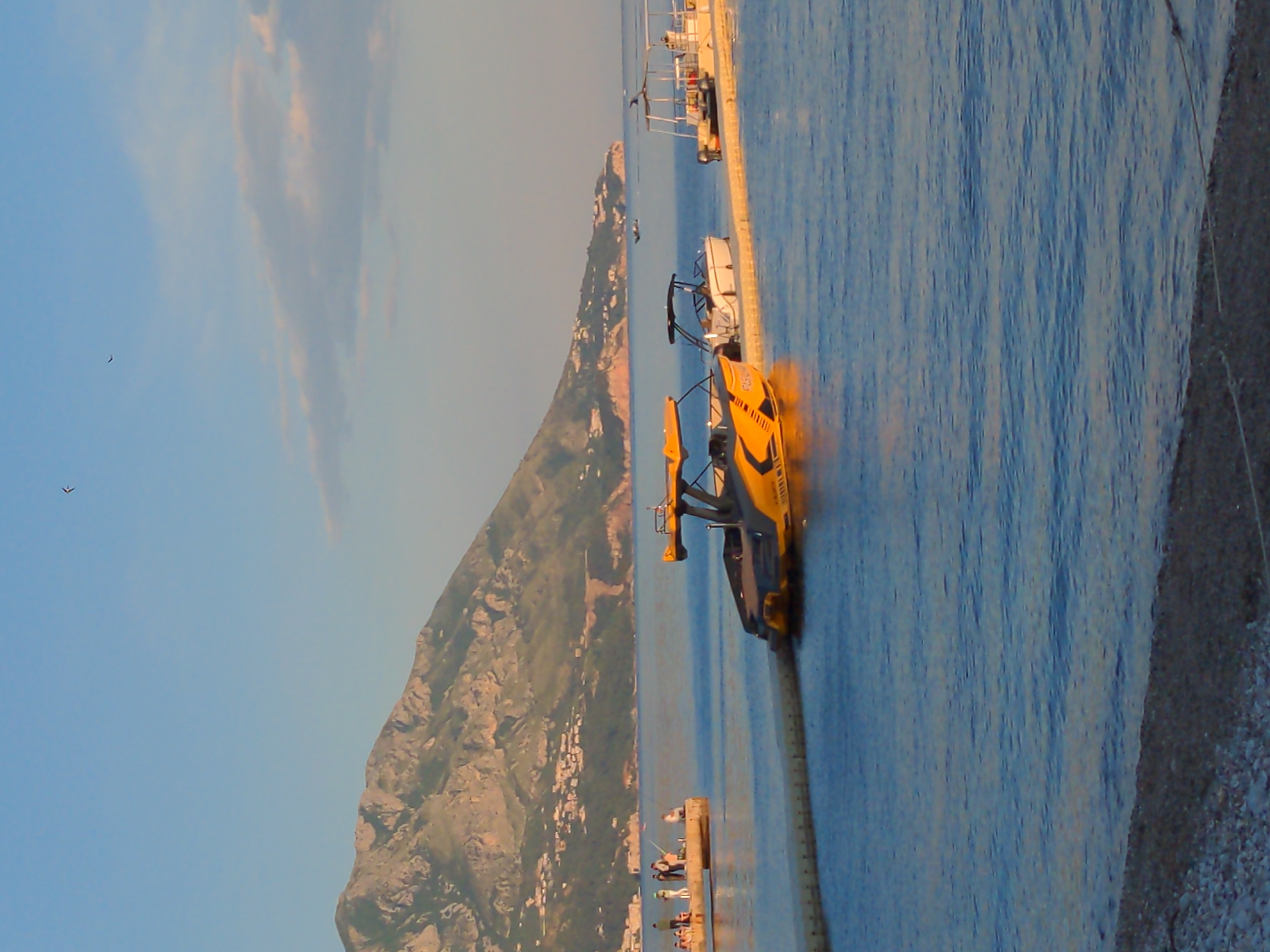 The image depicts a sunny coastal scene. A yellow and black boat is docked next to a pier on the right side of the frame. In the background, there is a tall mountain with houses and buildings nestled along its slopes. A group of people can be seen standing on a nearby dock. The sky is clear with light clouds. The sea appears calm.