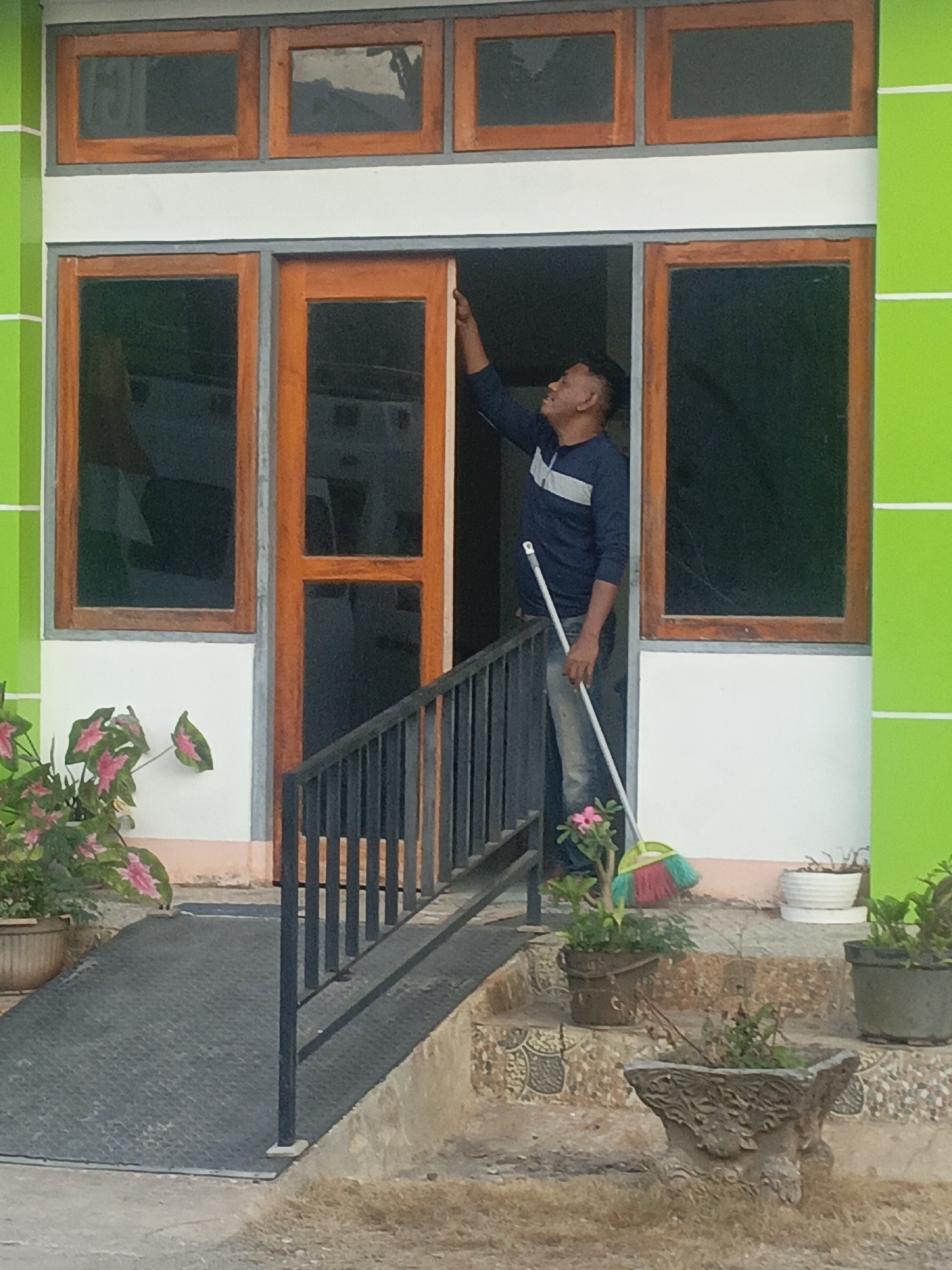 A man wearing a dark blue long-sleeved shirt with a white stripe across the chest and light-colored jeans stands in a doorway of a building with green and white walls. He is holding a broom with a multi-colored head and a long white handle, and appears to be sweeping. The doorway has wooden trim. In front of the building, there is a ramp with a black railing and stairs with patterned tiles leading up to the entrance. Several potted plants are visible on the ramp and stairs. The background shows windows with wooden frames. The scene is outdoors, with dry grass visible in the foreground.