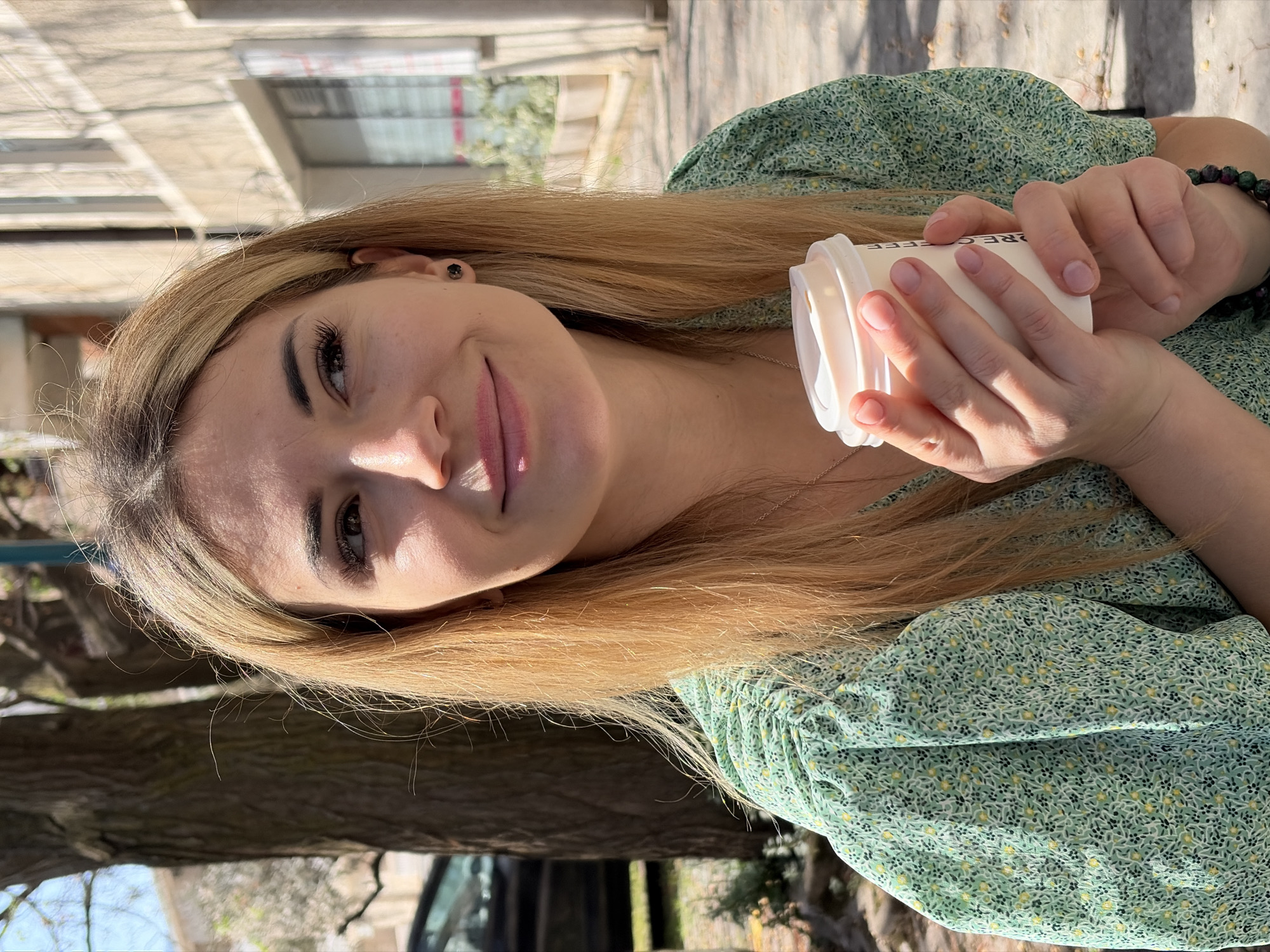 The image displays a young woman outdoors, holding a white coffee cup. She has long, straight blonde hair and is wearing a light green dress adorned with a small, intricate floral pattern. Her head is tilted slightly, and she looks towards the camera with a gentle, relaxed smile, suggesting a pleasant and comfortable mood.
Her hands are clasped around the coffee cup, which has black text partially visible, appearing to read "RE COFFEE" or "ORE COFFEE." On her right wrist, she wears a bracelet made of dark, possibly reddish-green, beaded stones.
The background reveals a sunny outdoor setting, likely a park or urban street. To the left of the woman, a light-colored building with visible windows is present, partially shadowed. To her right, a large tree trunk with textured bark is prominent. Further in the distance, glimpses of a street or path and what might be a parked vehicle are visible under bright, natural daylight. The strong sunlight casts soft shadows, indicating it is daytime. The overall scene conveys a casual and serene moment. The location context is Budva, Montenegro.