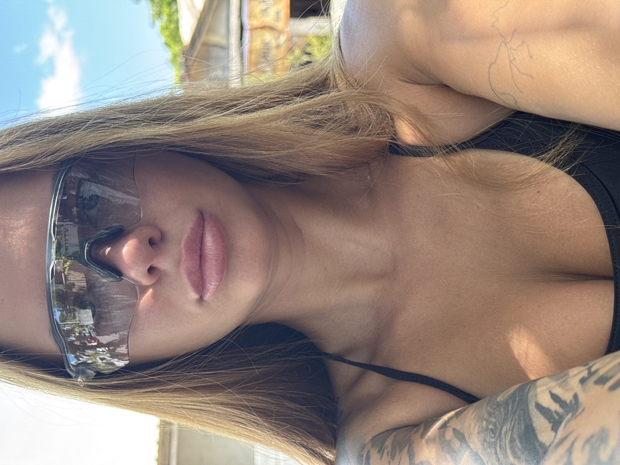 A selfie of a person with long blond hair wearing sunglasses and a black top is shown in the image. The person has a tattoo on their right arm and another on their left shoulder. The sunglasses reflect a scene with trees and buildings, suggesting an outdoor setting. The background is slightly blurred, but trees and buildings are visible. Based on the prompt, this image was taken in North Kuta, Indonesia.