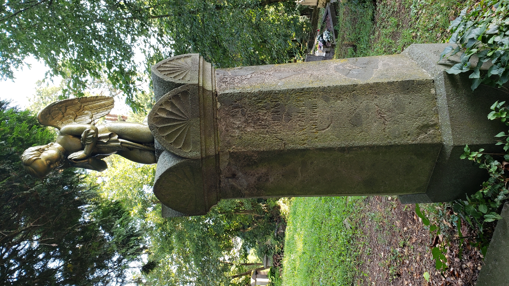 The image shows a gravestone in a cemetery, likely in the city of Prešov, Slovakia. A golden statue of a cherub is kneeling on top of the gravestone, with its hands clasped in prayer and wings outstretched. The gravestone itself is made of stone and has inscriptions that are partially obscured by moss and weathering. The surrounding environment is a cemetery with grass, trees, and other gravestones visible in the background. The lighting suggests it is daytime, with sunlight filtering through the trees. The overall scene is somber and reflective, common in a cemetery setting.