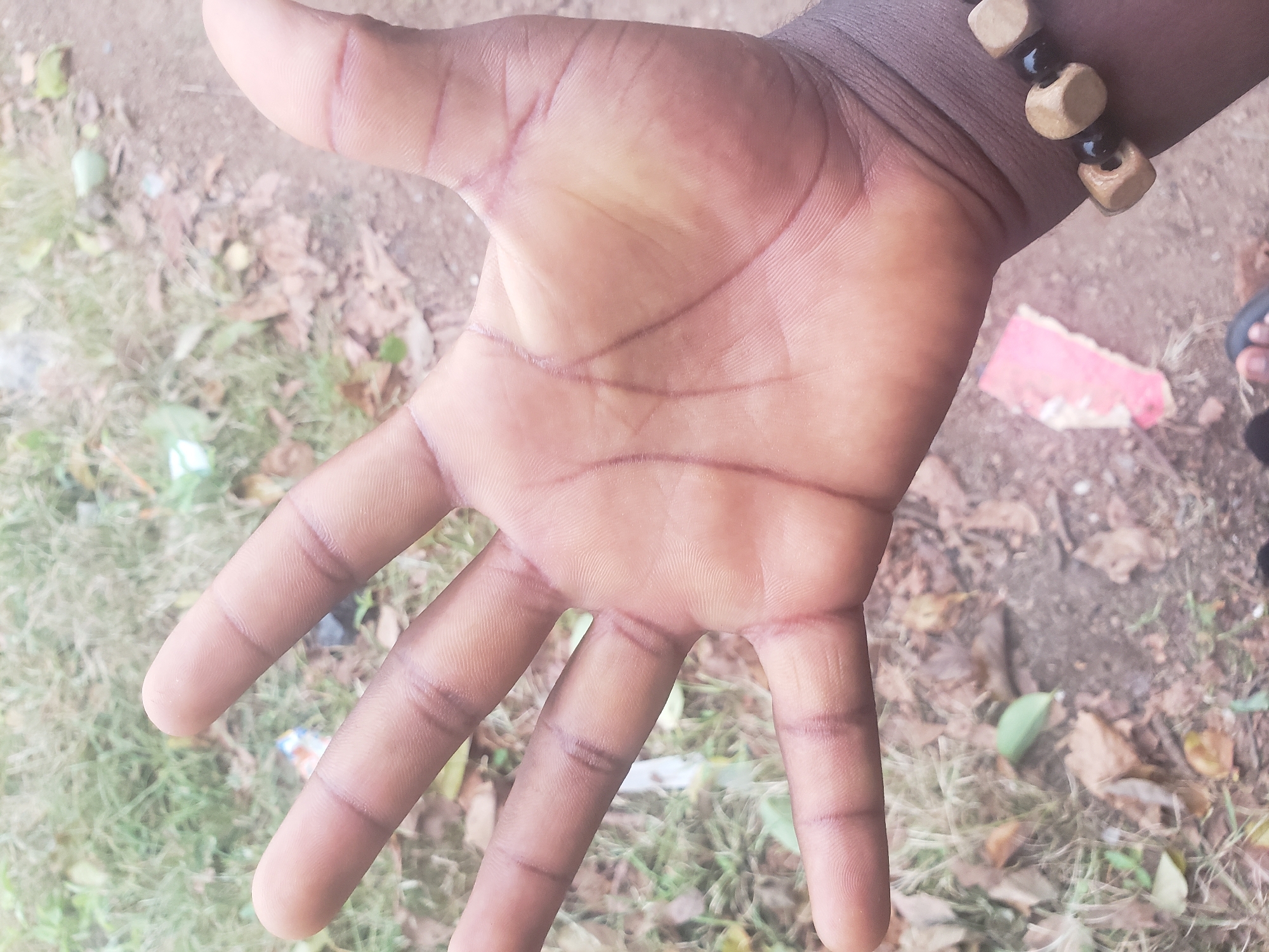 The image displays a close-up view of a left human hand, palm facing upwards, against a blurry outdoor background. The hand is tanned with visible palm lines and creases on the fingers, which are spread open. A beaded bracelet, composed of wooden cubes and black beads, is worn on the wrist. The background consists of dry leaves, dirt, and some sparse green vegetation, suggesting a natural or unkempt outdoor environment. There are no identifiable landmarks or contextual clues to determine a specific location within a city.
