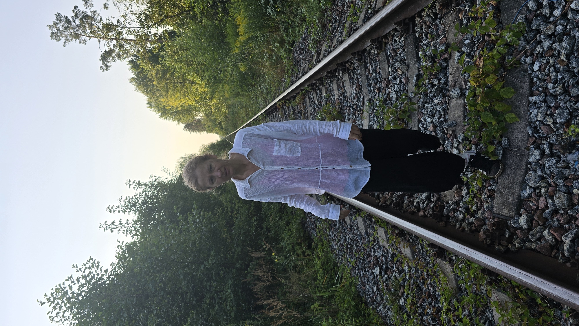 A medium shot shows a woman walking down a set of train tracks in a wooded area. The woman is positioned in the center of the frame, facing the camera. She has blonde hair pulled back and is wearing a white button-down shirt over a pink garment, dark pants, and black sneakers. Her expression is neutral. The train tracks run diagonally from the foreground into the distance, flanked by dense green foliage and trees. The ground is covered in gravel and scattered vegetation. The sky above is clear and pale, suggesting it is either early morning or late evening. The scene appears to be located in a rural or semi-rural area, possibly on the outskirts of Stanyshivka, Ukraine, where overgrown train lines are not uncommon.