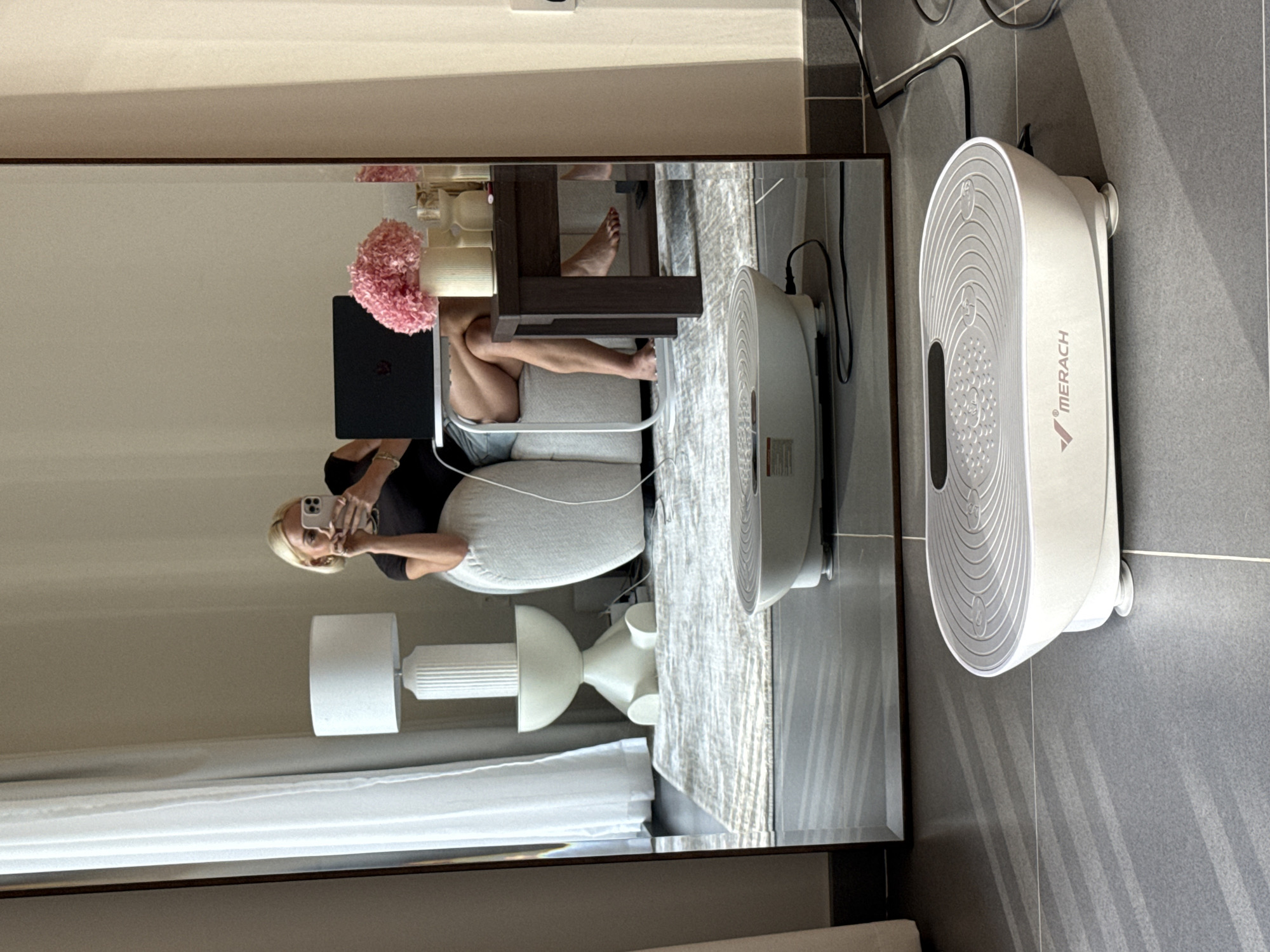 The image, taken as a mirror selfie, depicts an indoor scene in what appears to be a modern living area or bedroom. The setting is provided as Dubai, United Arab Emirates.
In the reflection of a large, dark-framed mirror, a blonde woman is visible. She is wearing a dark t-shirt and holding a smartphone up to take the picture, with her face and neutral expression visible. She is seated on a light-colored upholstered sofa or chair, with her legs bent and one foot propped up. Beside her on a dark wooden table are a black laptop, a pink floral arrangement, and some white decorative items. Further back in the reflection, two white lamps – one with a classic shade and another with a sculptural base – are visible, along with white curtains and a light-colored, textured rug on the floor.
Outside the mirror's reflection, on a gray tiled floor, a white, oval-shaped electronic device is prominently displayed. The device has a ribbed, textured surface and the brand name "MERACH" is clearly visible on its side. It features small feet and is connected by black electrical cables that run along the gray tiled wall. The lighting suggests it is daytime, with shadows cast on the tiled wall from the device. The overall scene conveys a moment of casual activity or relaxation.
