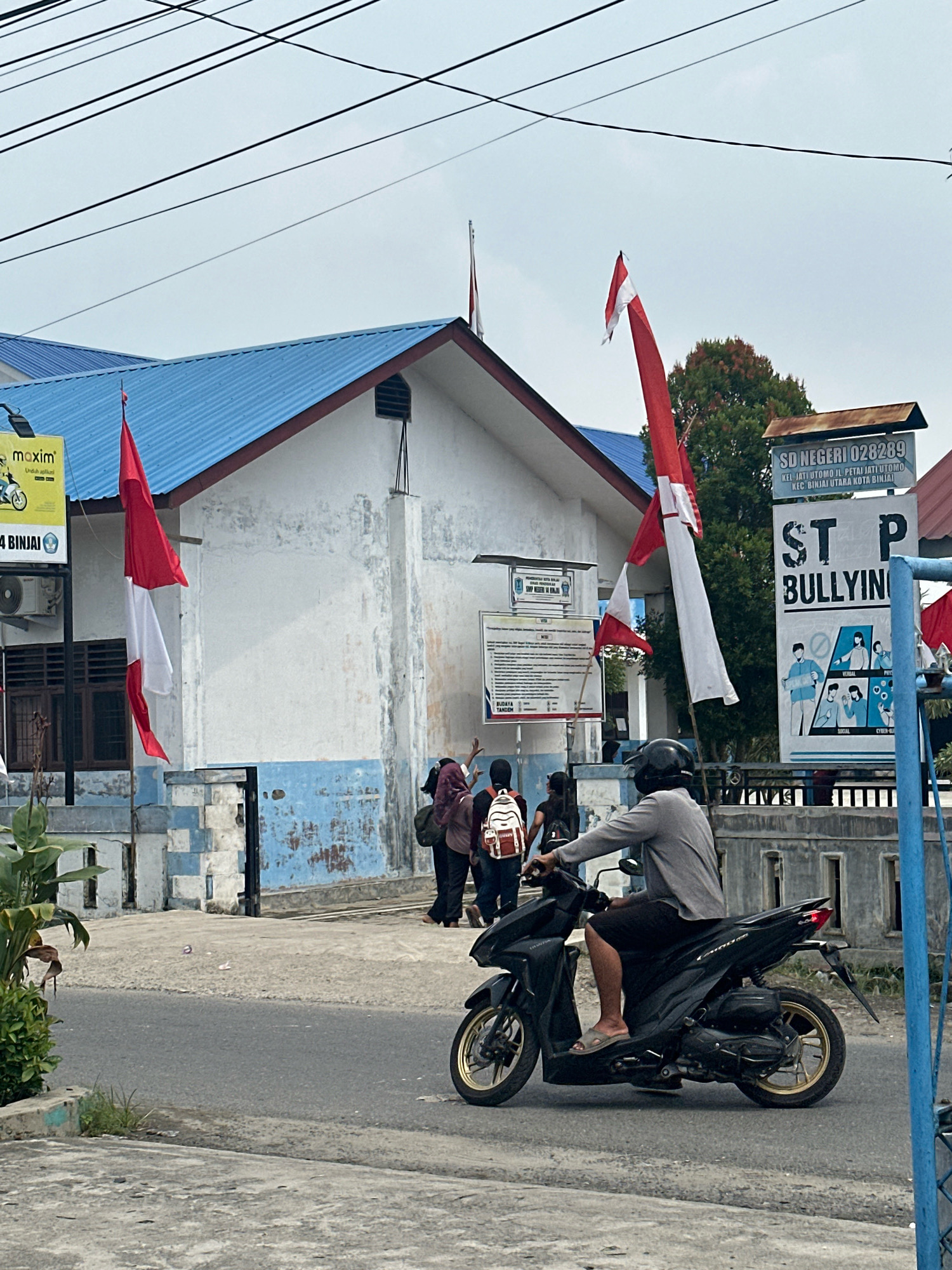 In the City of Binjai, Indonesia, a street scene unfolds near a school building with a blue roof. A person rides a black motorcycle, their legs exposed, wearing a grey long-sleeved shirt, black shorts, and sandals. The motorcycle has gold rims. Behind the motorcyclist, a group of individuals walks on a paved area in front of the school. One person carries a white backpack with red accents. The school building exhibits a white facade with blue lower trim. Several Indonesian flags, red and white, are prominently displayed, some fluttering from poles. A large sign on the right reads "ST P BULLYING" with accompanying illustrations depicting different forms of bullying. Another sign to the left of the "ST P BULLYING" sign displays information for "SD NEGERI 028289" (State Elementary School 028289), including its address in Jati Utomo and Binjai Utara. A smaller yellow sign with the "maxim" logo is also visible on the left side of the building. Overhead, a network of black electrical wires crisscrosses the sky. The ground is paved with asphalt and concrete. Green foliage is present in the lower left corner.