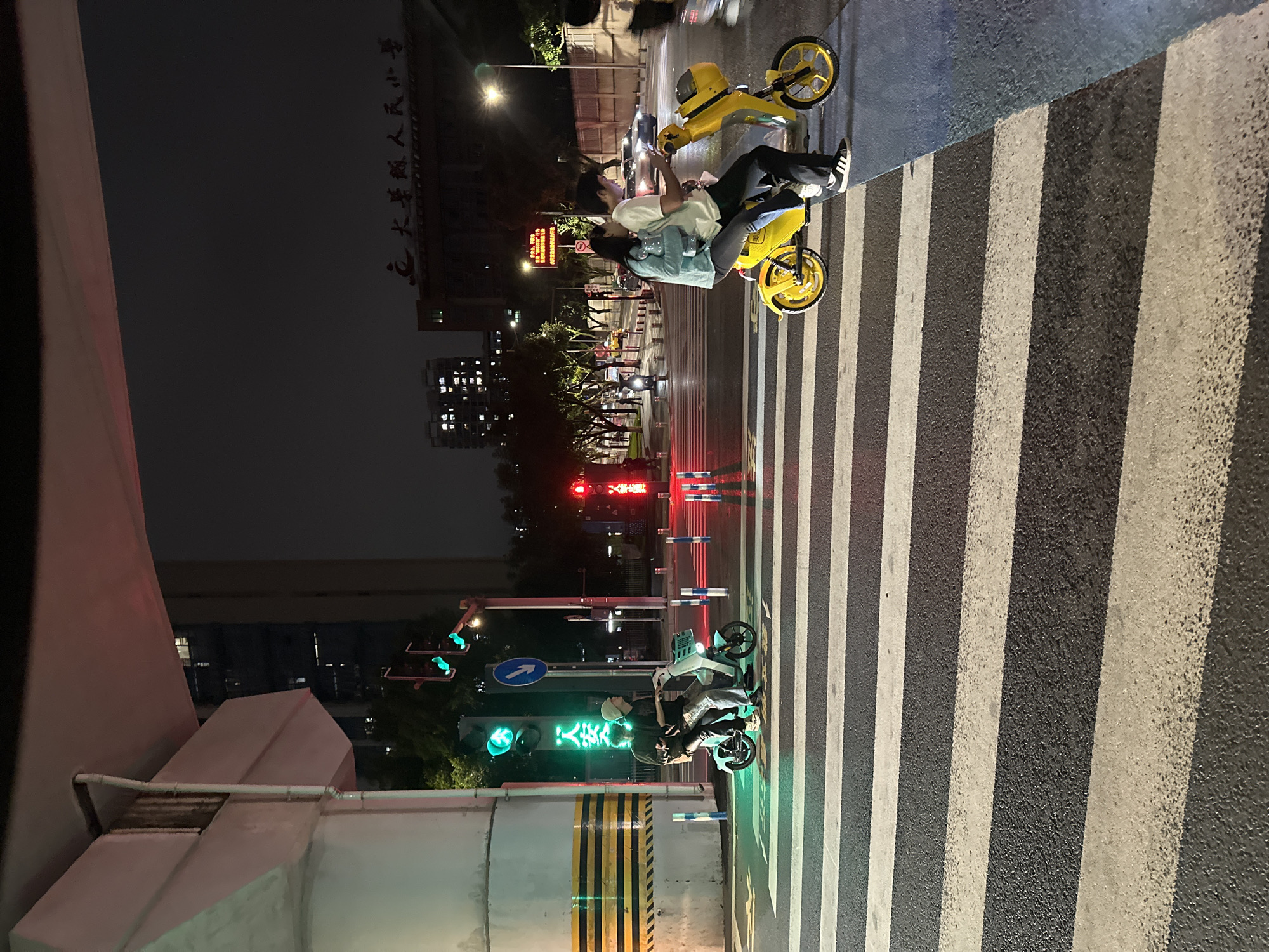 At night, two people ride an electric scooter on a crosswalk in the Chongqing High-tech Zone, China. The crosswalk is wet, reflecting the green and red lights of a traffic signal. A second, lighter colored electric scooter is also visible on the crosswalk, carrying two people. In the background, buildings and streetlights illuminate the urban environment. The overall scene depicts a typical nighttime streetscape in a Chinese city.