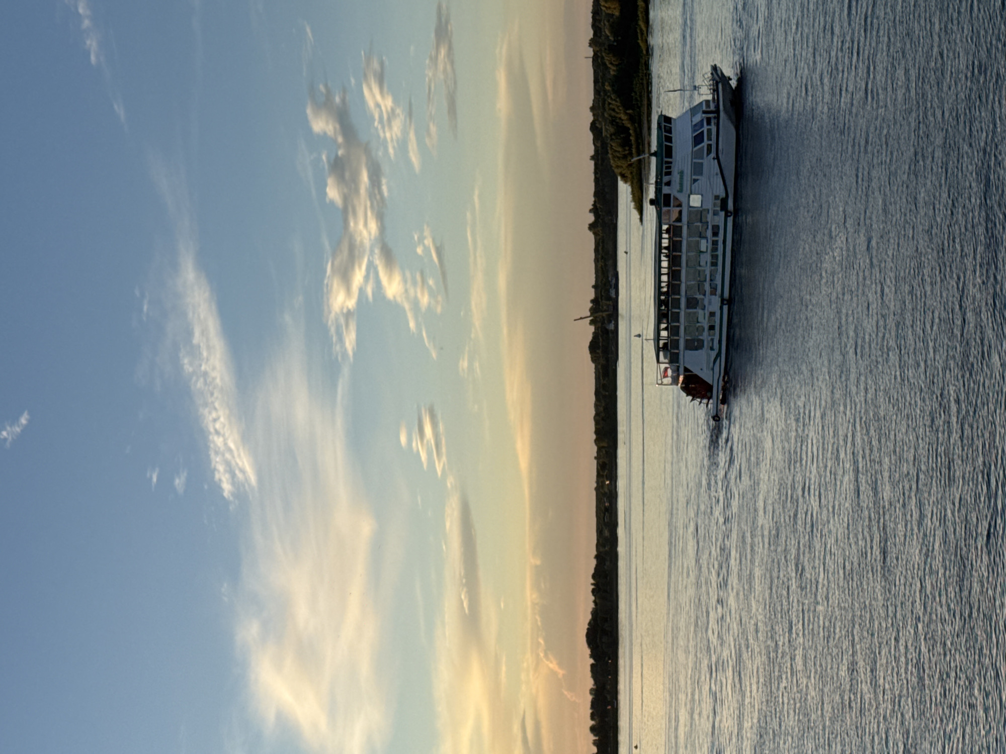 The image depicts a cruise ship on a wide river, likely the Volga River, with a tree-lined bank in the background. The sky is a vibrant mix of blue with streaks of white and orange clouds, suggesting either sunrise or sunset. The water is calm with subtle ripples. The ship, painted white with green accents, has multiple decks and windows, indicating it is carrying passengers. The overall scene conveys a peaceful and scenic river cruise experience. The location is likely Nizhny Novgorod, Russia, a city situated at the confluence of the Oka and Volga rivers, known for its historical significance and picturesque riverfront.