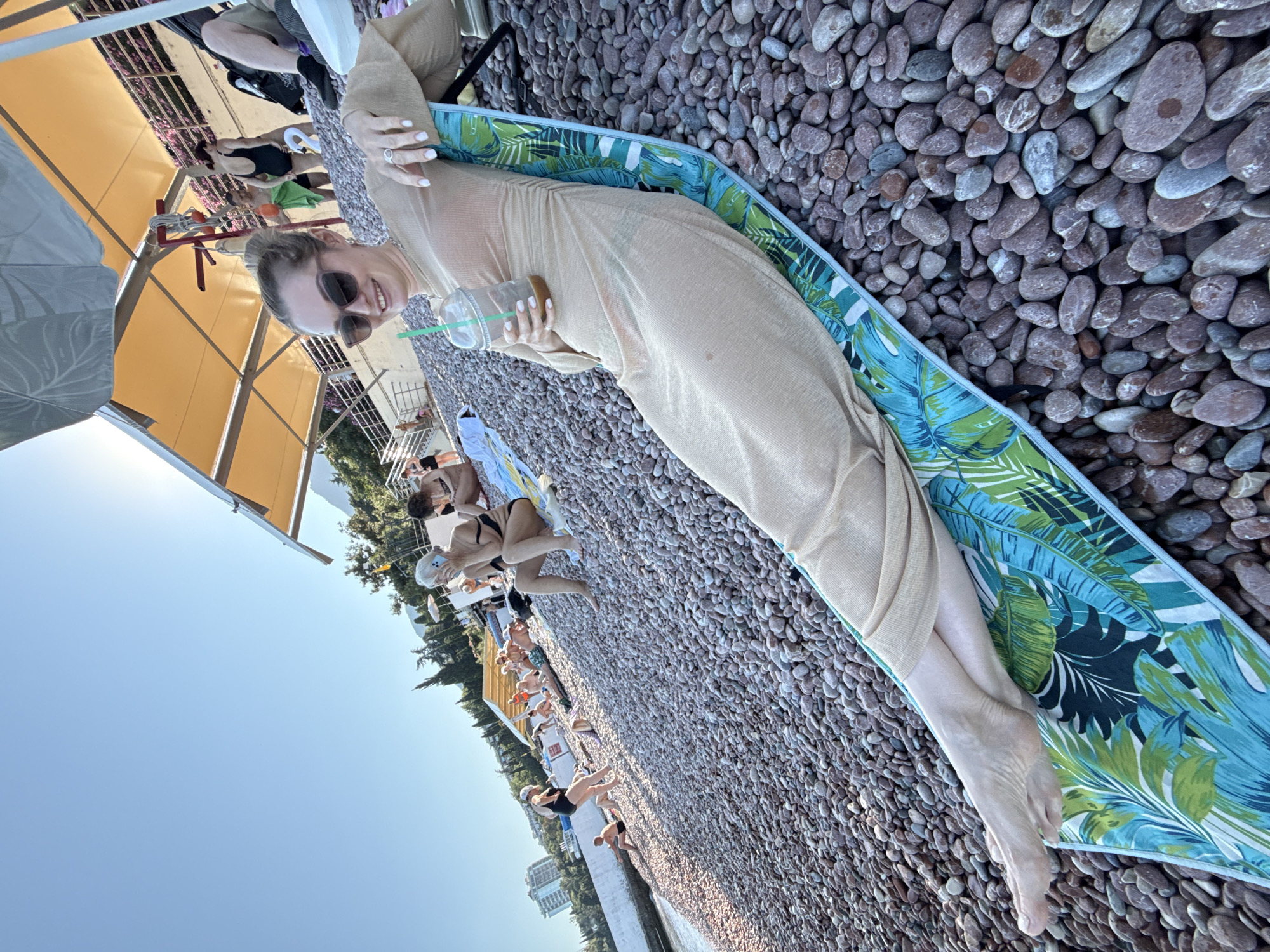 This is a high-angle, medium shot of a woman relaxing on a beach in Gurzuf, Ukraine. The woman is lying on a beach towel with a tropical leaf print, on a pebbled beach. She is wearing a sheer, beige, long-sleeved dress and sunglasses, and holding a plastic cup with a straw. Her legs are crossed, and her feet are resting on the towel. In the background, several other people are visible on the beach, some lying on towels and others walking. A yellow awning shades part of the beach, and trees and buildings are visible on the hillside behind. The sky is clear and blue.