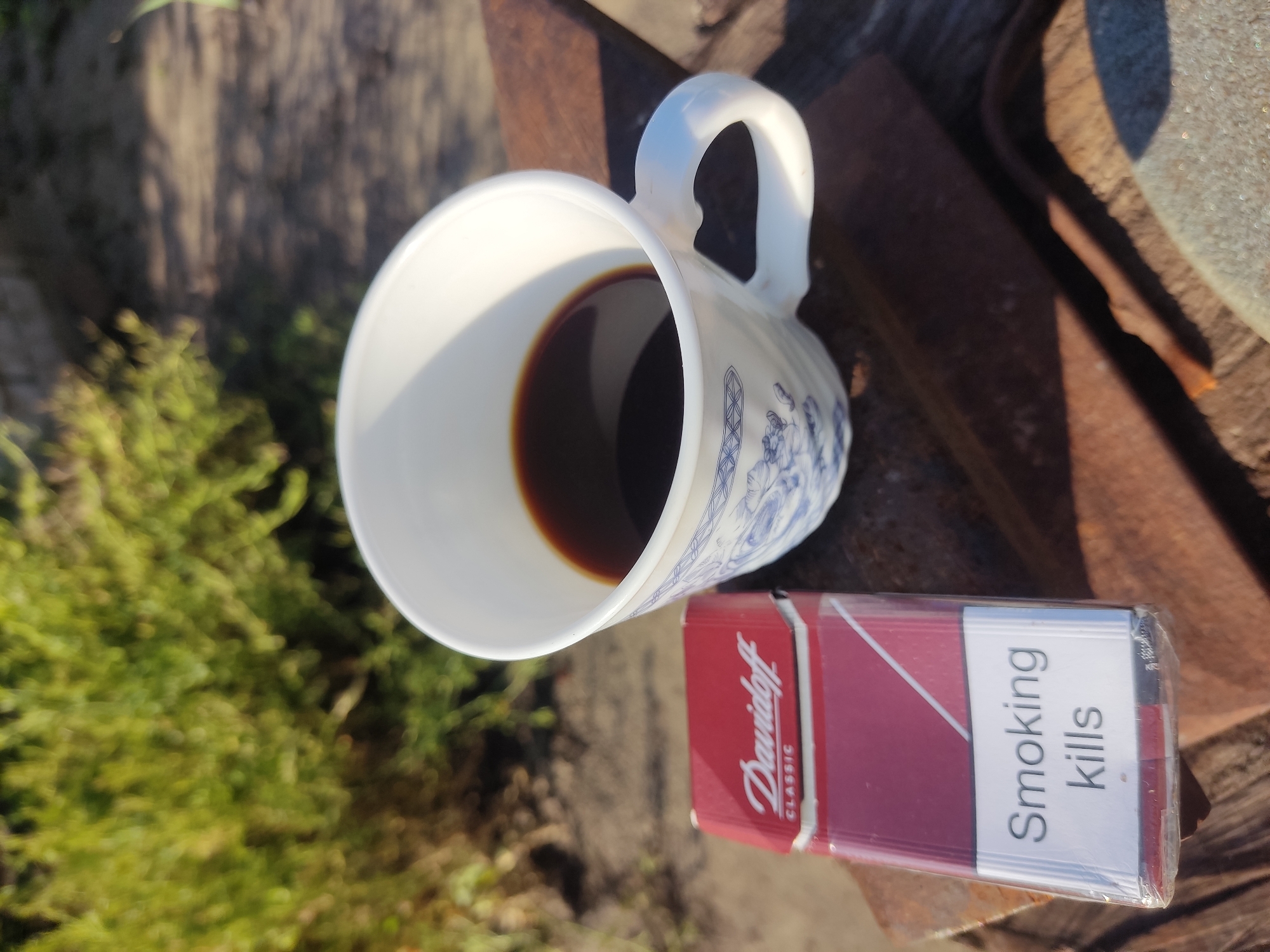 A pack of Davidoff Classic cigarettes and a white mug filled with dark liquid, likely coffee, are placed on a rusty metal surface outdoors. The mug has blue intricate designs on its exterior. The background is blurred and appears to be foliage, suggesting an outdoor setting. The cigarette pack has a warning label that reads "Smoking kills." The location is identified as Radisne, Ukraine.
