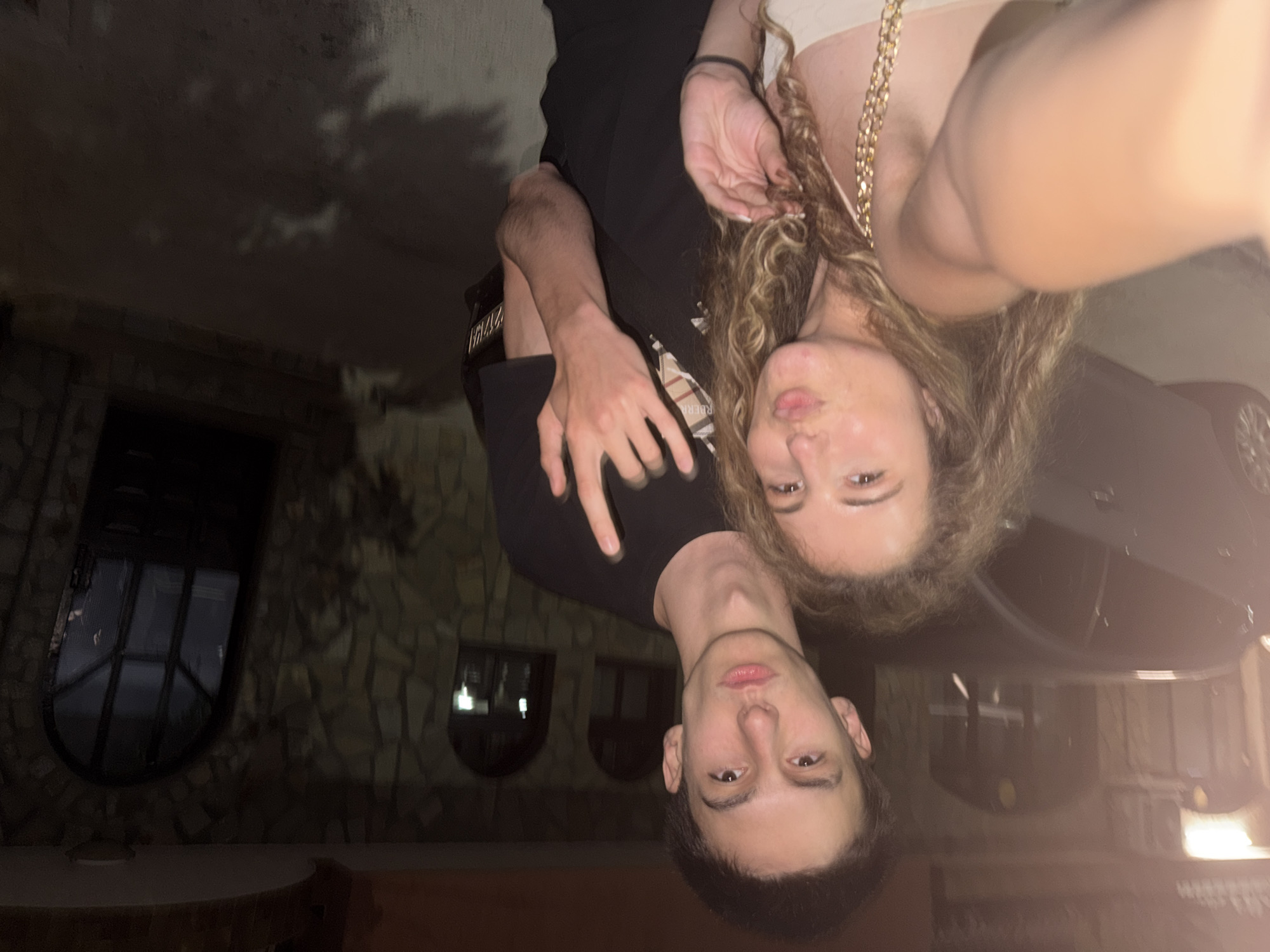 The image is a selfie taken by two young people at night, outdoors. The background shows a stone building with arched windows. A dark car is visible in the foreground. The lighting is dim, suggesting it is nighttime. The man in the foreground is wearing a black t-shirt and has short dark hair. The woman next to him has curly blonde hair and is wearing a light-colored top. They are both looking towards the camera. The orientation of the image is sideways. The location is the city of Lyubimets, Bulgaria.